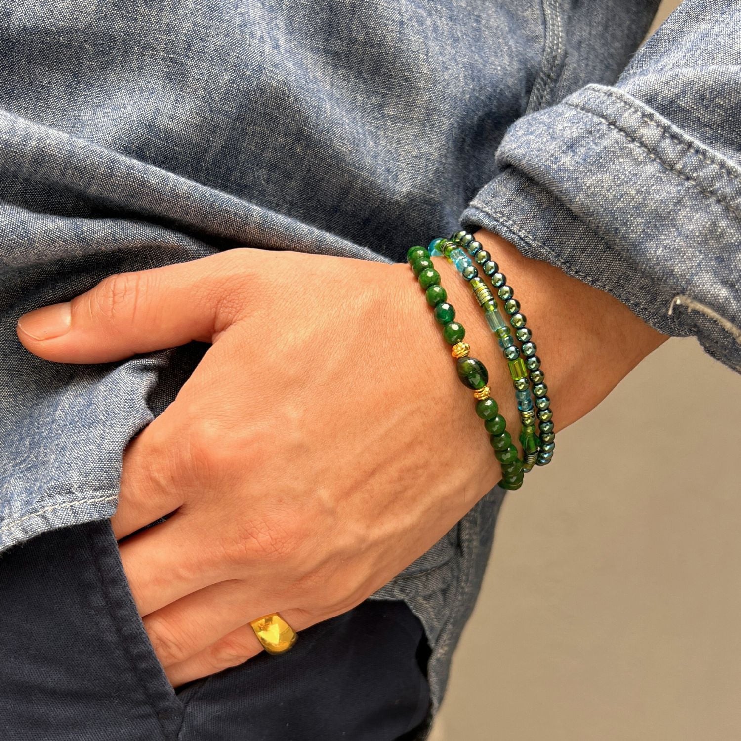 Wearing a denim shirt, a person slips a hand into their pocket, displaying the JAREDJAMIN Jewelry Online Emerald City Bracelet Set—green hematite and gold stretch bracelets—paired with a chunky gold pinky ring against a plain backdrop.
