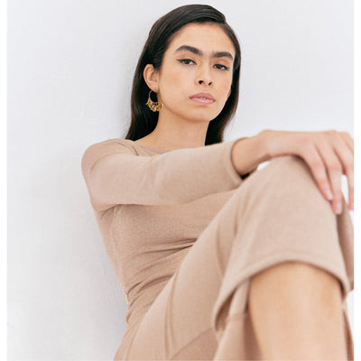 A woman with long dark hair wears a beige, long-sleeved outfit and JAREDJAMIN Jewelry Online’s Gold Large Petali Click-Top Hoop Earrings. She sits against a white background, one knee raised, exuding confidence in a minimal, modern setting.