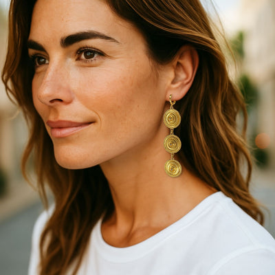A woman with wavy brown hair in a white shirt smiles slightly, wearing JAREDJAMIN Jewelry Online’s Gold Minerva Leverback Earrings—18K gold-plated with three ornate circular patterns. A softly blurred background suggests an outdoor setting.