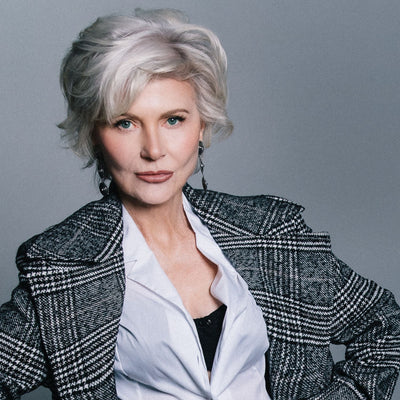 An older woman with short white hair poses confidently in bold makeup, a black-and-white plaid jacket, and white blouse. She wears Minerva Black Clip on Earrings from JAREDJAMIN Jewelry Online, standing out against a plain gray background.