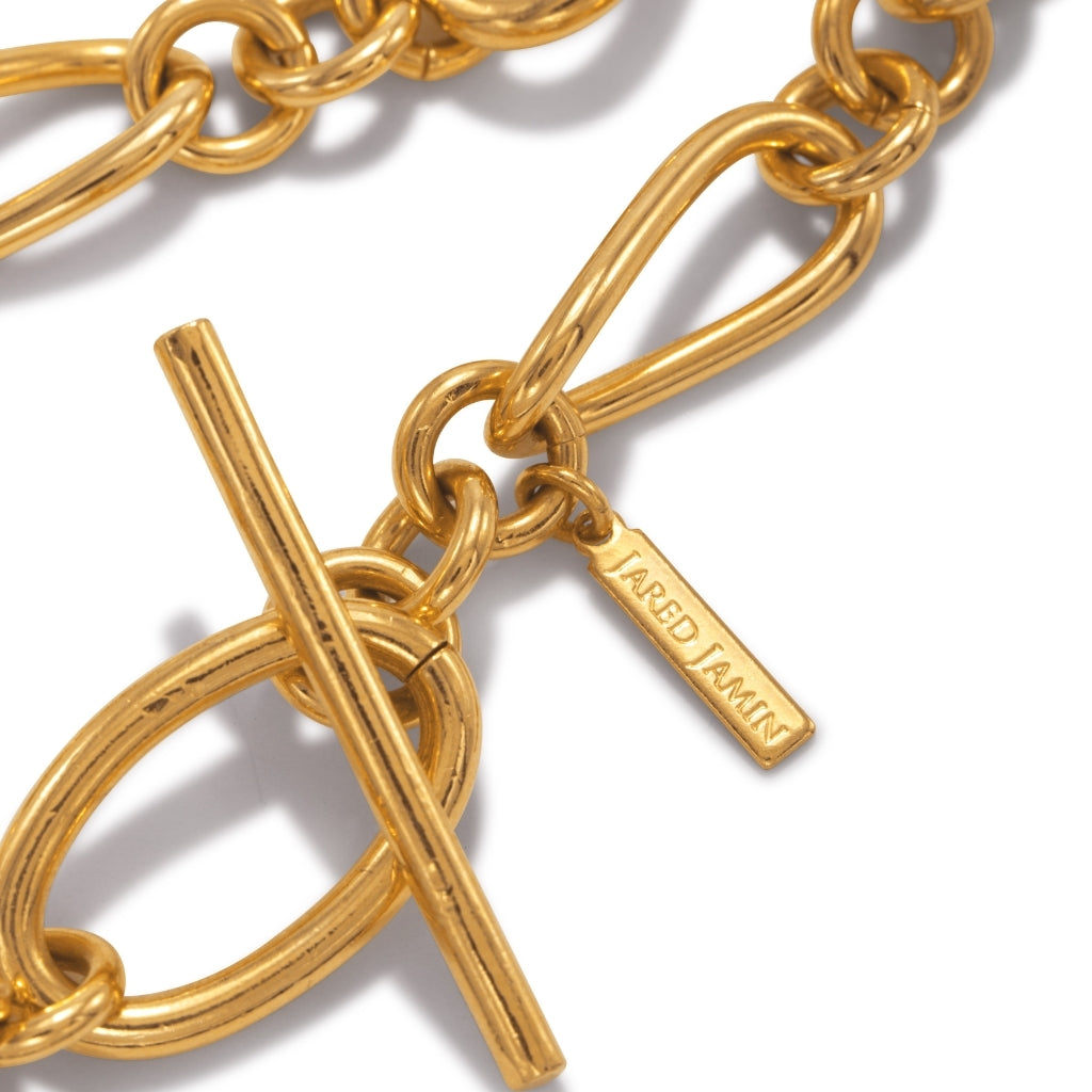 Close-up of a gold chain with a toggle clasp. A small rectangular tag with engraved text is attached to one link. The image highlights the chain's shiny surface, robust links, and elegant design.