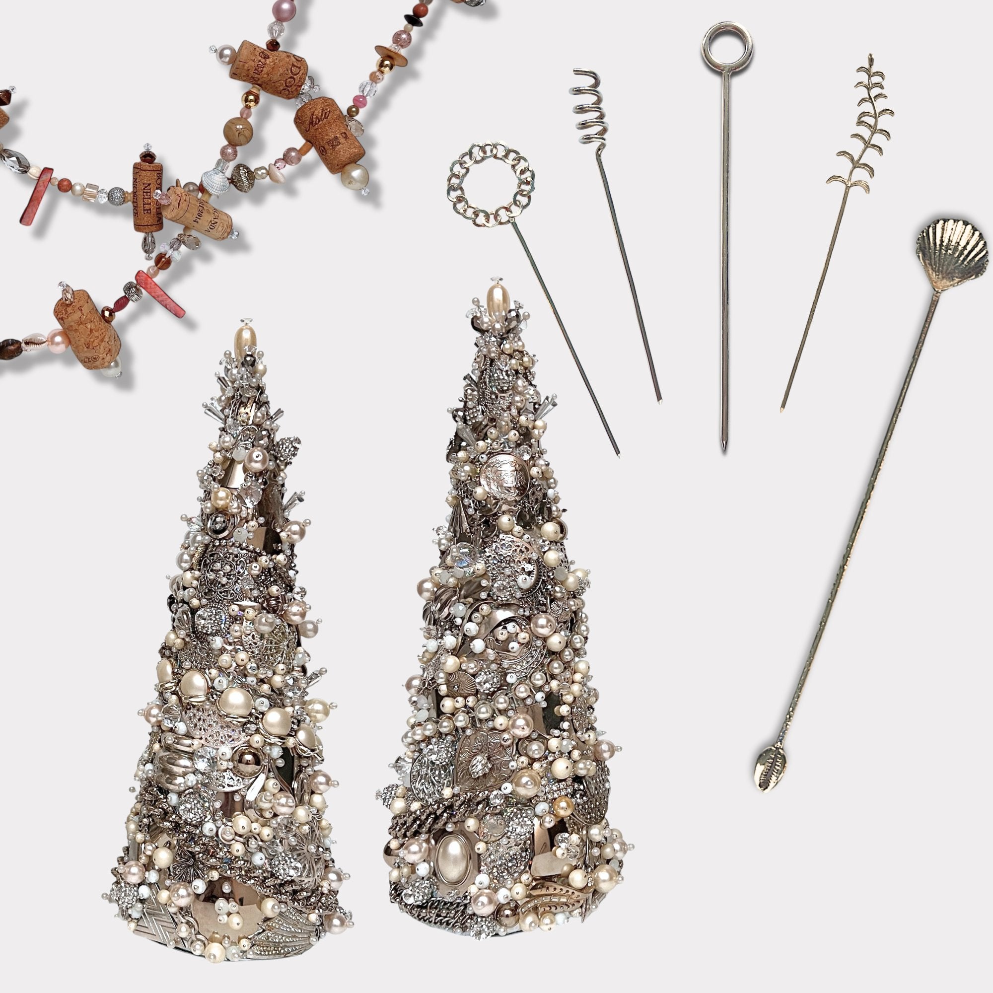 Two cone-shaped sculptures adorned with pearls and jewelry, surrounded by decorative metal skewers and a whimsical drink stirrer with a shell on one end, lay near a wine-themed beaded necklace.