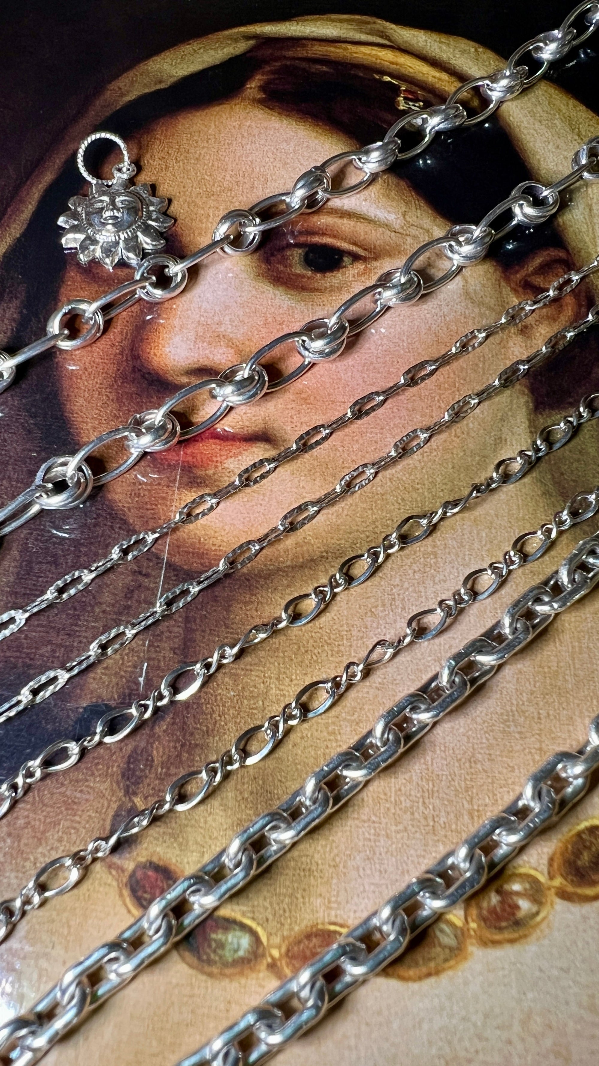 Several silver chains and a floral pendant are arranged diagonally across a vintage painting of a woman’s face, partially covering her features and contrasting with the artwork’s soft tones.