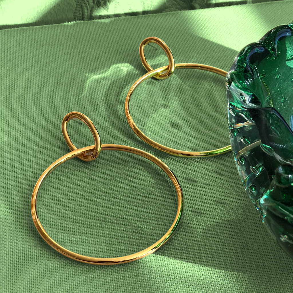 Gold hoop earrings on a textured green surface beside a reflective glass object, with soft shadows creating a serene ambiance.