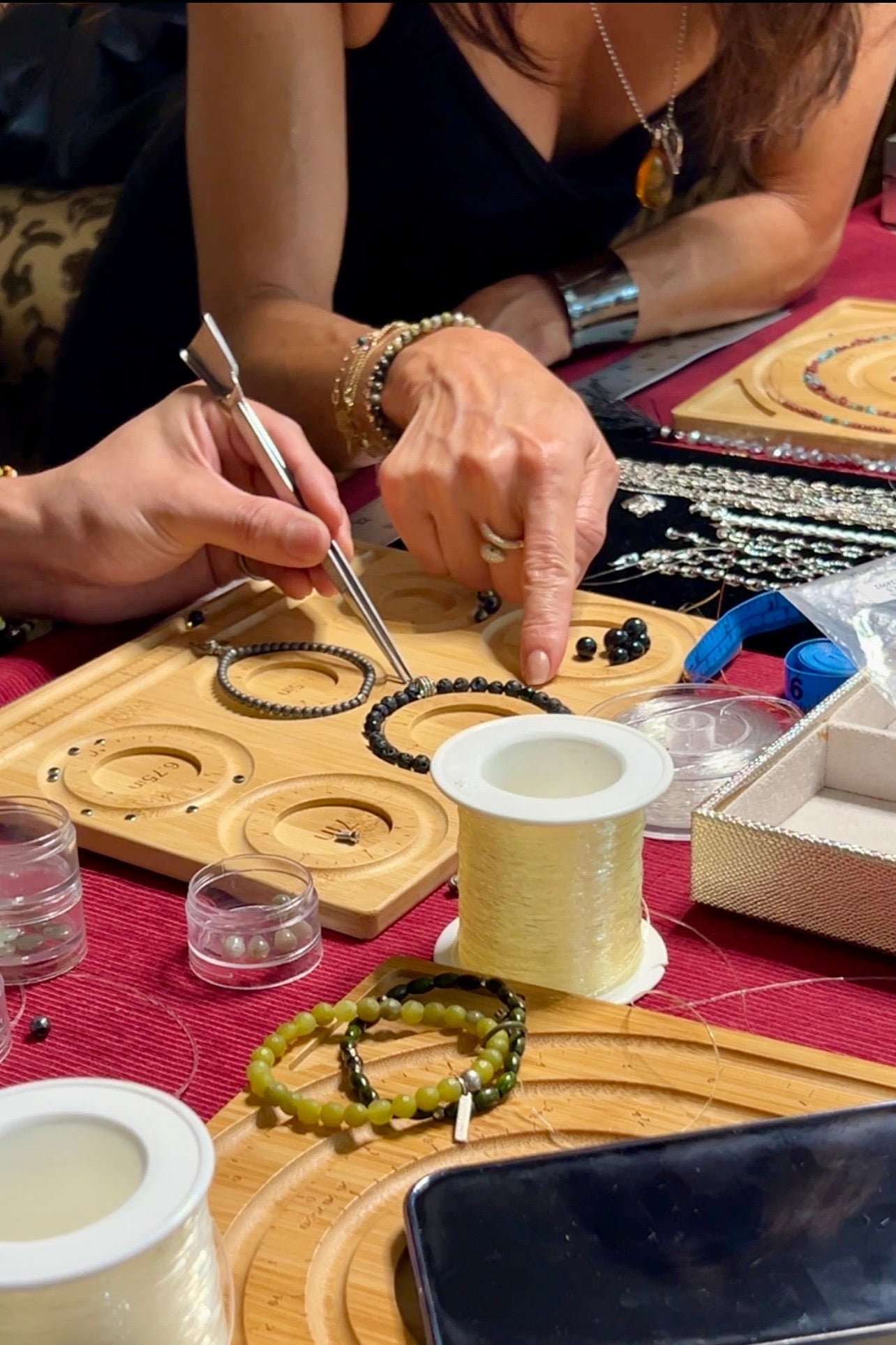 Two women make beaded jewelry at a table covered in tools and supplies. One points at black beads with a tool while the other points with her finger. Spools of string and finished bracelets are visible.
