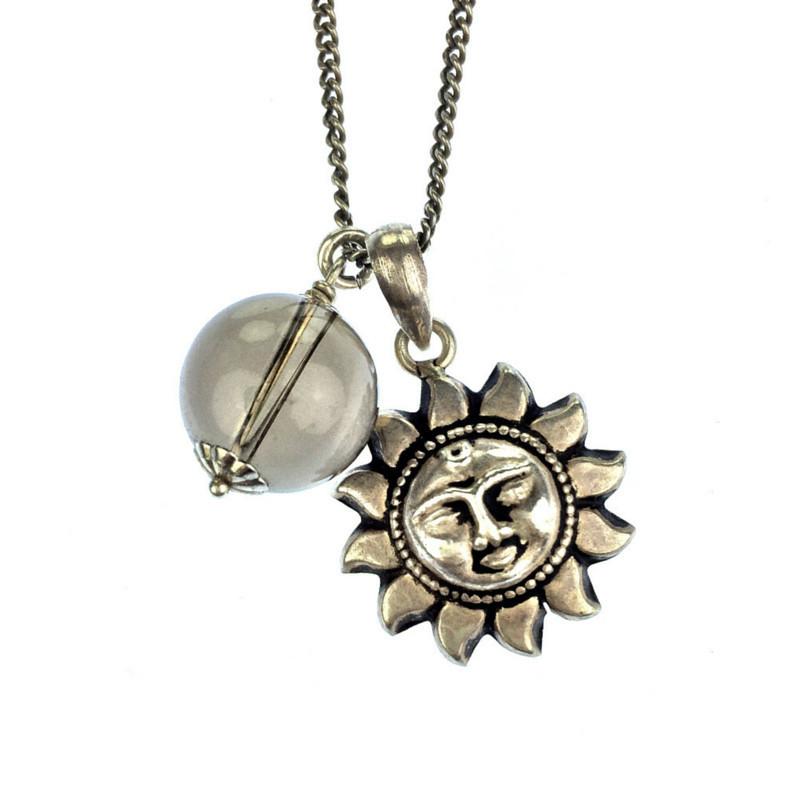 A pendant necklace with a silver sun charm featuring a face and a clear, spherical bead. The charms hang on a delicate silver chain against a white background.