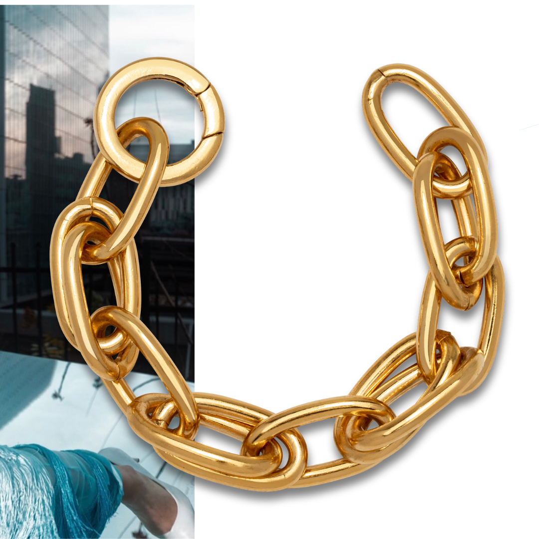 A gold chain bracelet with large oval links arranged in a semicircle. The background partially shows a cityscape with tall buildings and a person in a blue outfit.