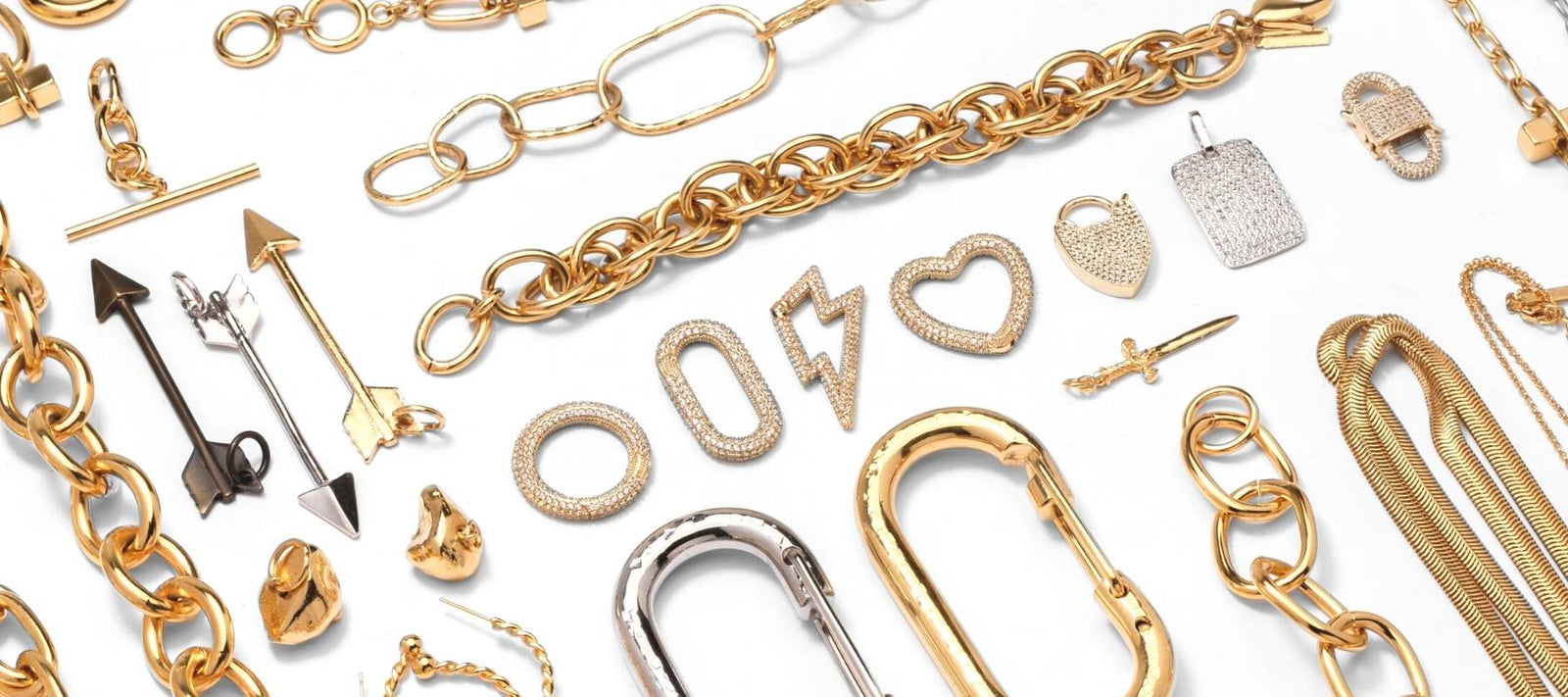 A collection of gold and silver jewelry on a white background, featuring various chains, earrings, pendants, and charms. Designs include arrows, a nail, a lightning bolt, a heart, padlocks, and various geometric shapes. The pieces are intricately detailed and diverse in style.