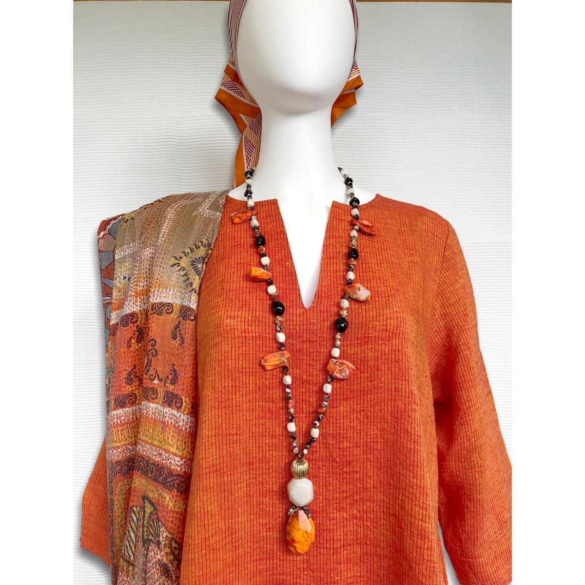 A mannequin models an orange V-neck tunic, JAREDJAMIN Firestarter ネックレスとorange stonesとjasper beads, a printed shawl over one shoulder,とa striped orange-and-ホワイト scarf wrapped around its head.