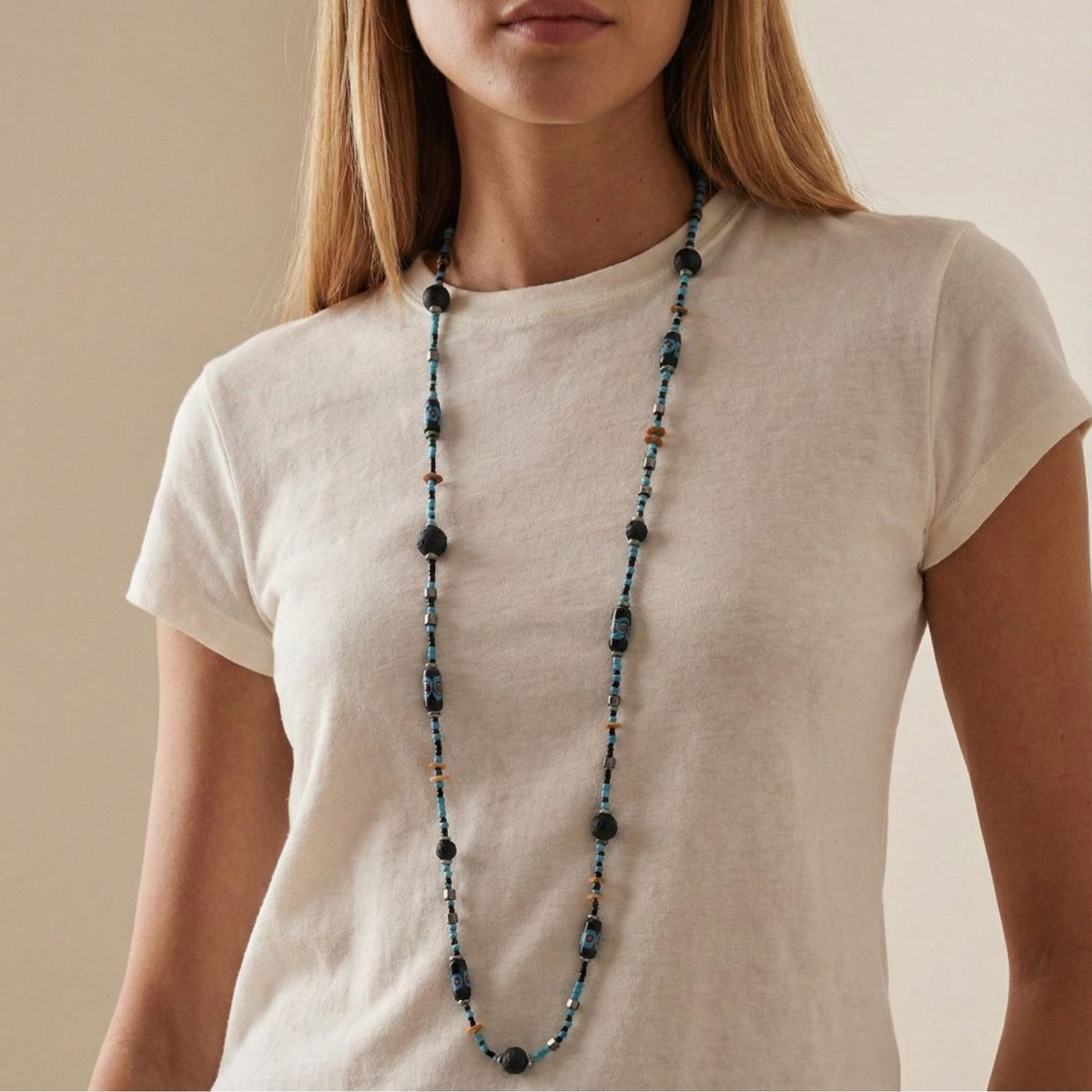 A woman with long blonde hair wears the JAREDJAMIN Jewelry Online African Voyage Long Lava Necklace, featuring dark blue, black, and copper beads, with a plain off-white t-shirt against a neutral background; her face is partially out of frame.