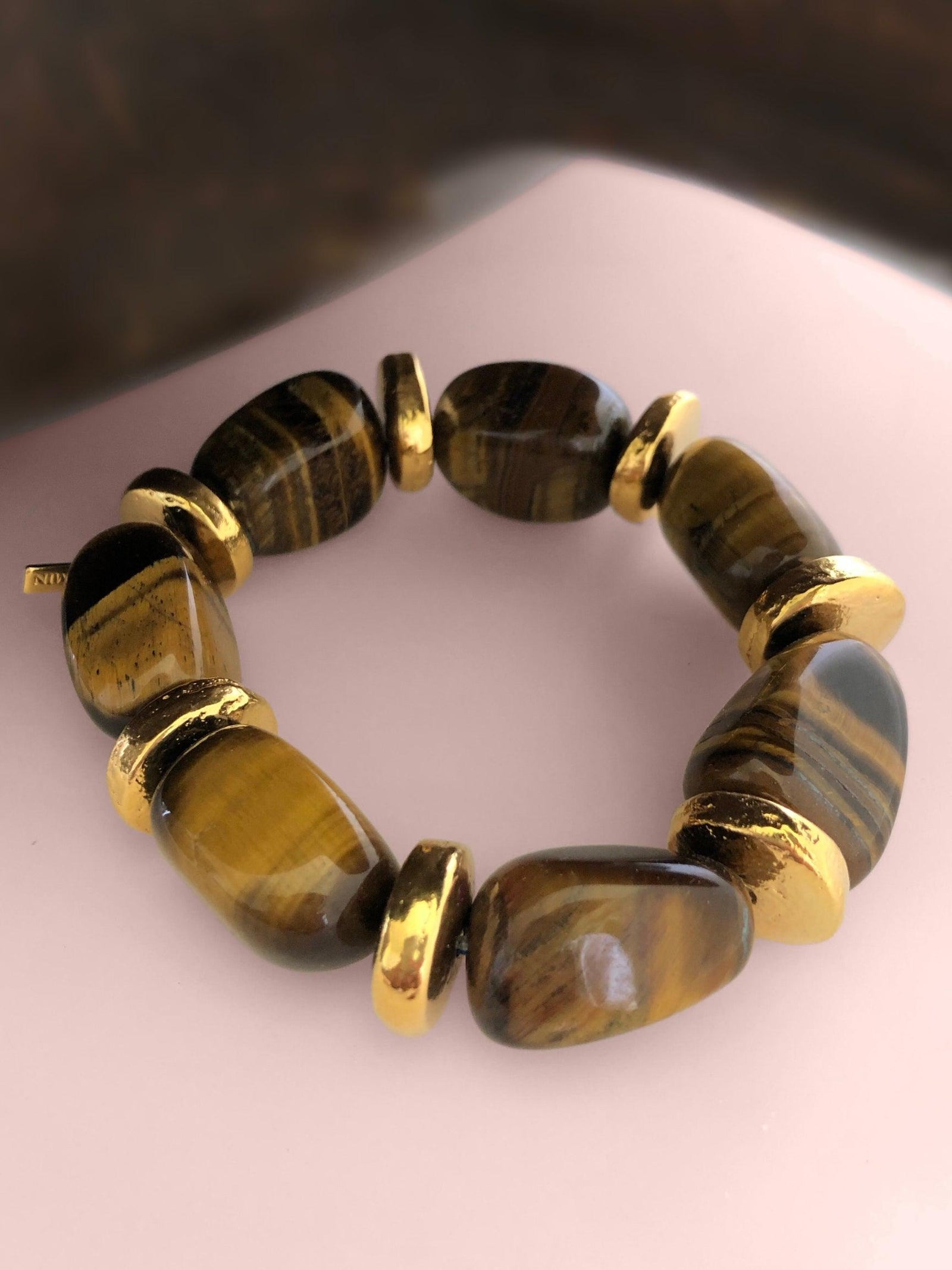 A bracelet made of large, polished tiger's eye stones and chunky gold spacer beads is arranged in a circle on a pale pink surface. The stones showcase striking brown and golden bands unique to tiger's eye jewelry.