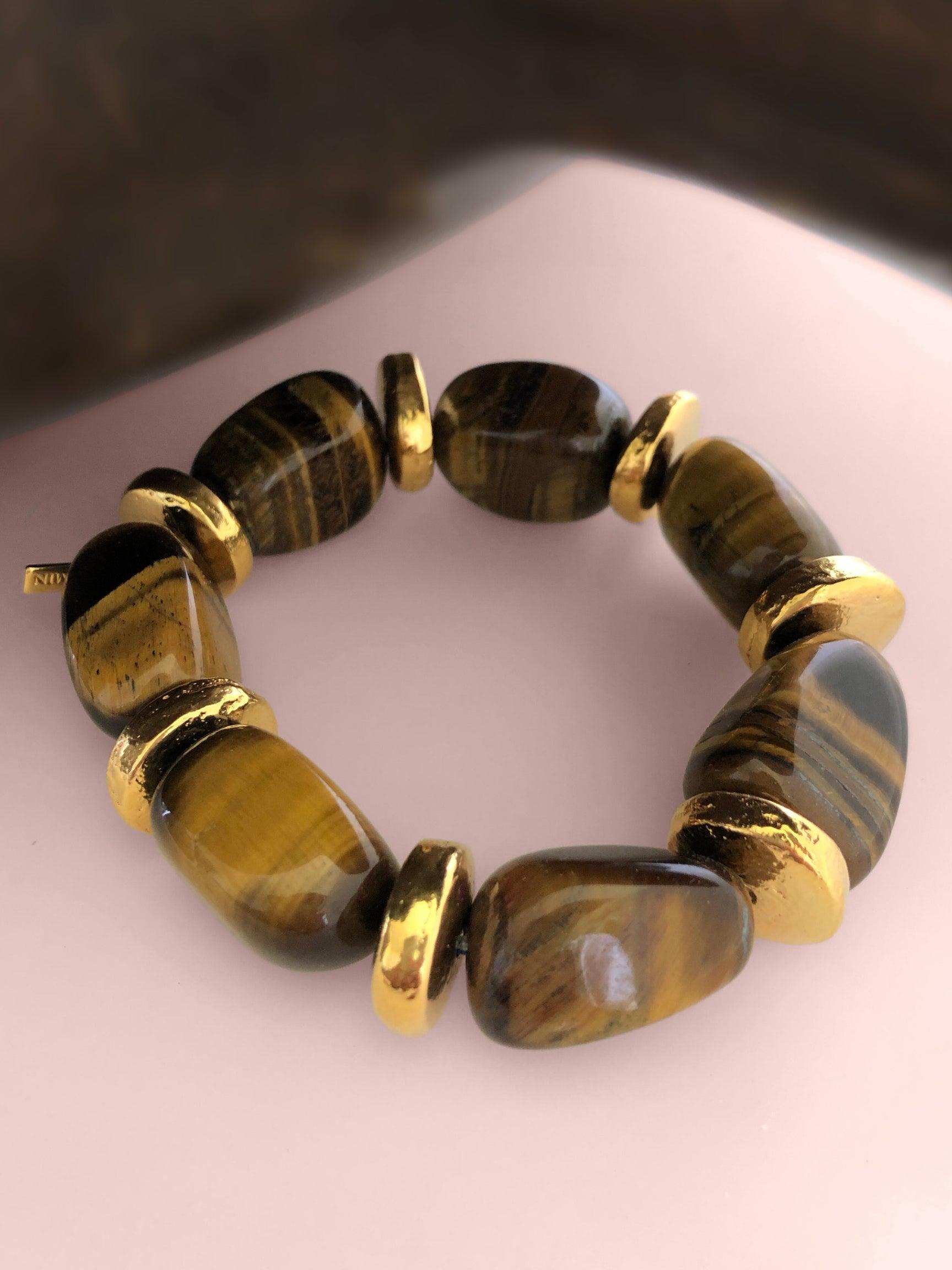A bracelet made of large, polished tiger's eye stones and chunky gold spacer beads is arranged in a circle on a pale pink surface. The stones showcase striking brown and golden bands unique to tiger's eye jewelry.