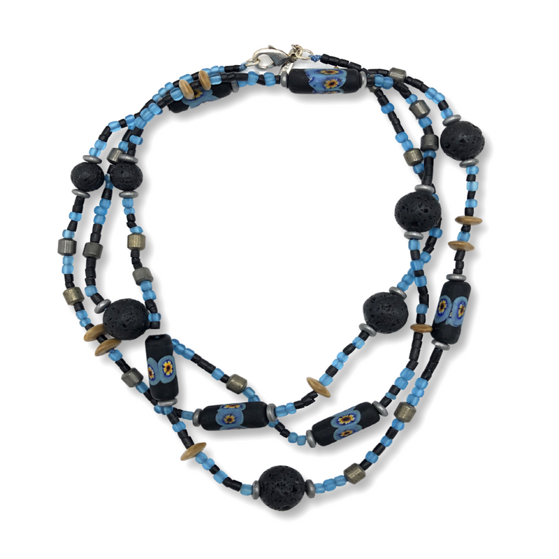 Coiled flat lay of the long beaded necklace on a white background, showing black lava rock rounds, black tube beads with blue and yellow floral patterns, turquoise and black seed beads, tan disc spacers, and silver-toned spacer beads, with the lobster clasp visible at the top.