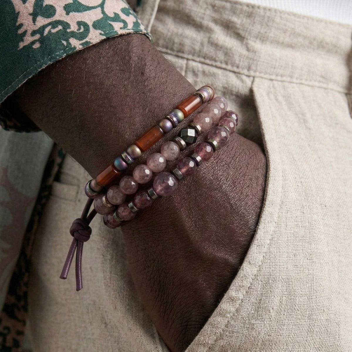 Detail shot of the Jared Jamin Sedona Dusk Bracelet Set styled with beige linen trousers. The image showcases the stack of faceted lavender gemstones, amber-toned glass, and rainbow hematite beads, emphasizing the rich textures and the signature purple leather cord tie.