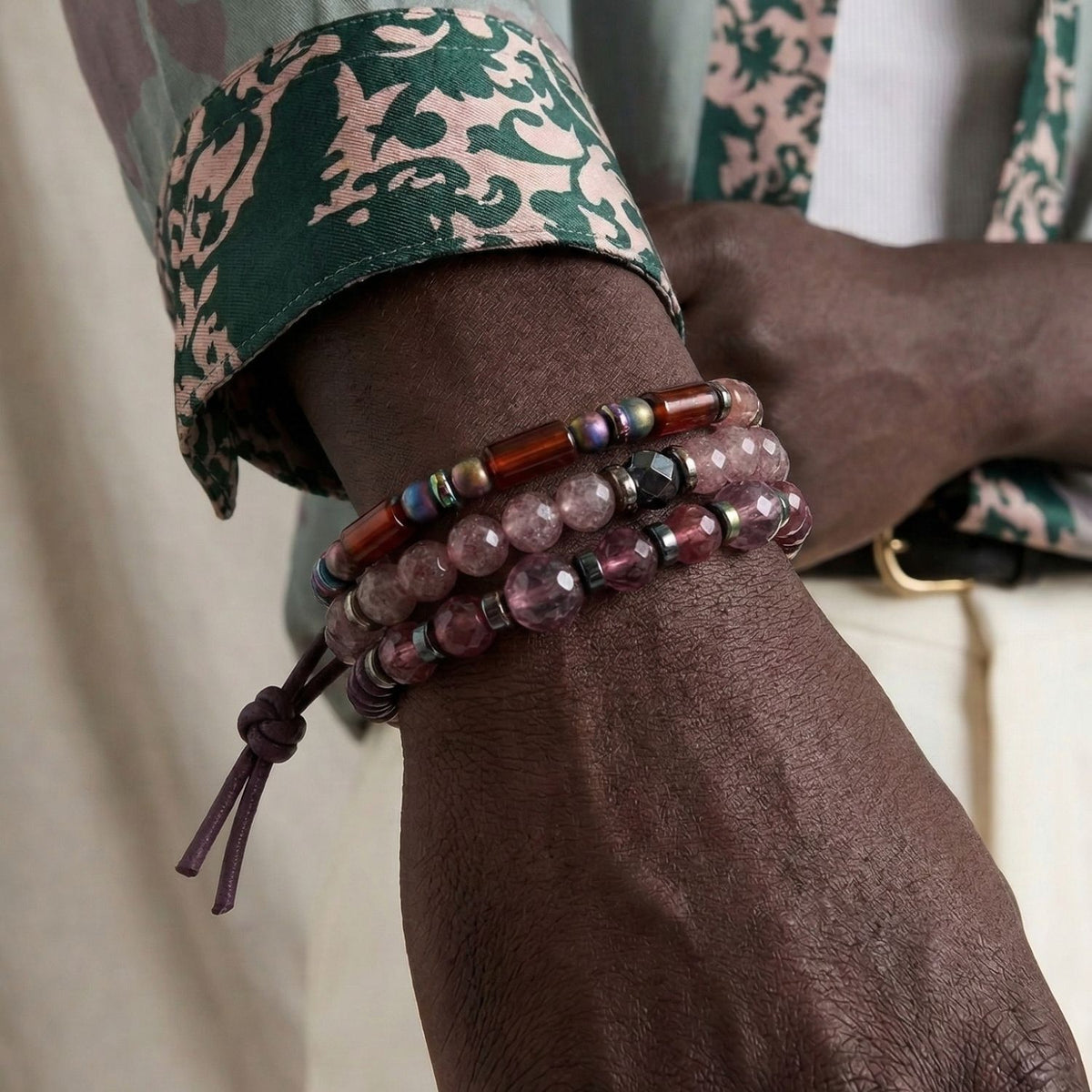 Men's designer jewelry styling featuring the Sedona Dusk Bracelet Set. The model wears a green and cream patterned silk shirt and white trousers, highlighting how the purple and earth-toned gemstones complement high-end summer fashion. Hand-assembled in West Hollywood.