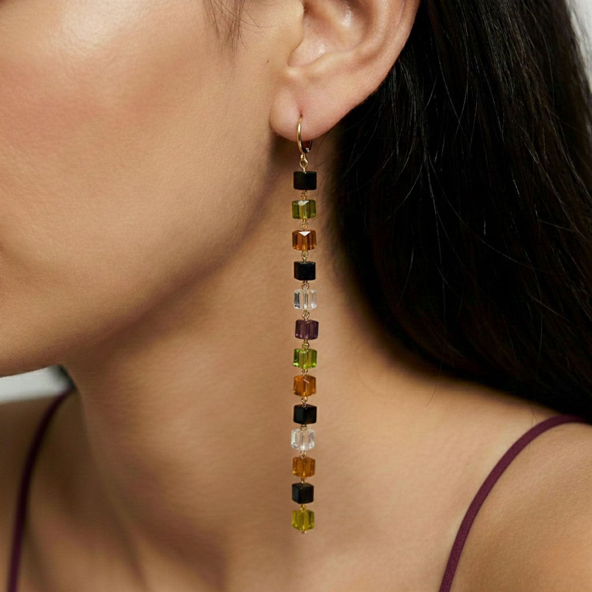 A close-up shows a woman's ear wearing Auburn Building Block Drop Earrings by JAREDJAMIN Jewelry Online, featuring stacked square beads in black, amber, clear, and green on 18K Gold Plated Lever Backs. She has straight dark hair and a burgundy top.