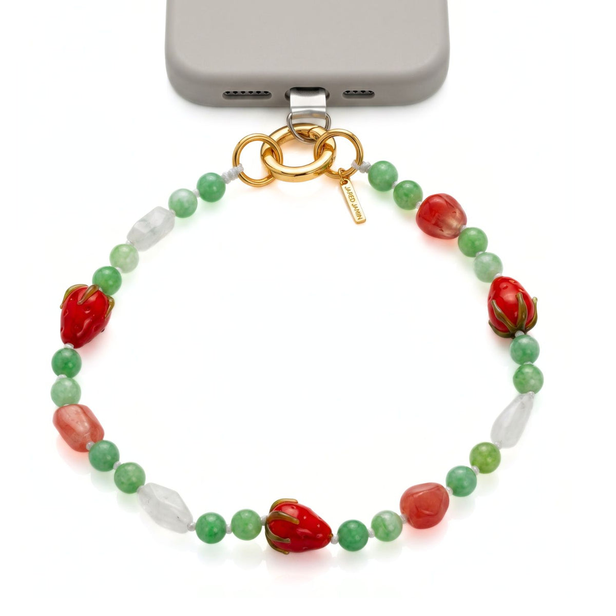 High-end studio shot of the JAREDJAMIN Berry Crush Phone Wristlet attached to a gray smartphone case via a clear anchor tab, showing the hand-knotted watermelon quartz, clear rock crystal, and glass strawberry beads connected to an 18k gold-plated circular clicker push clasp.