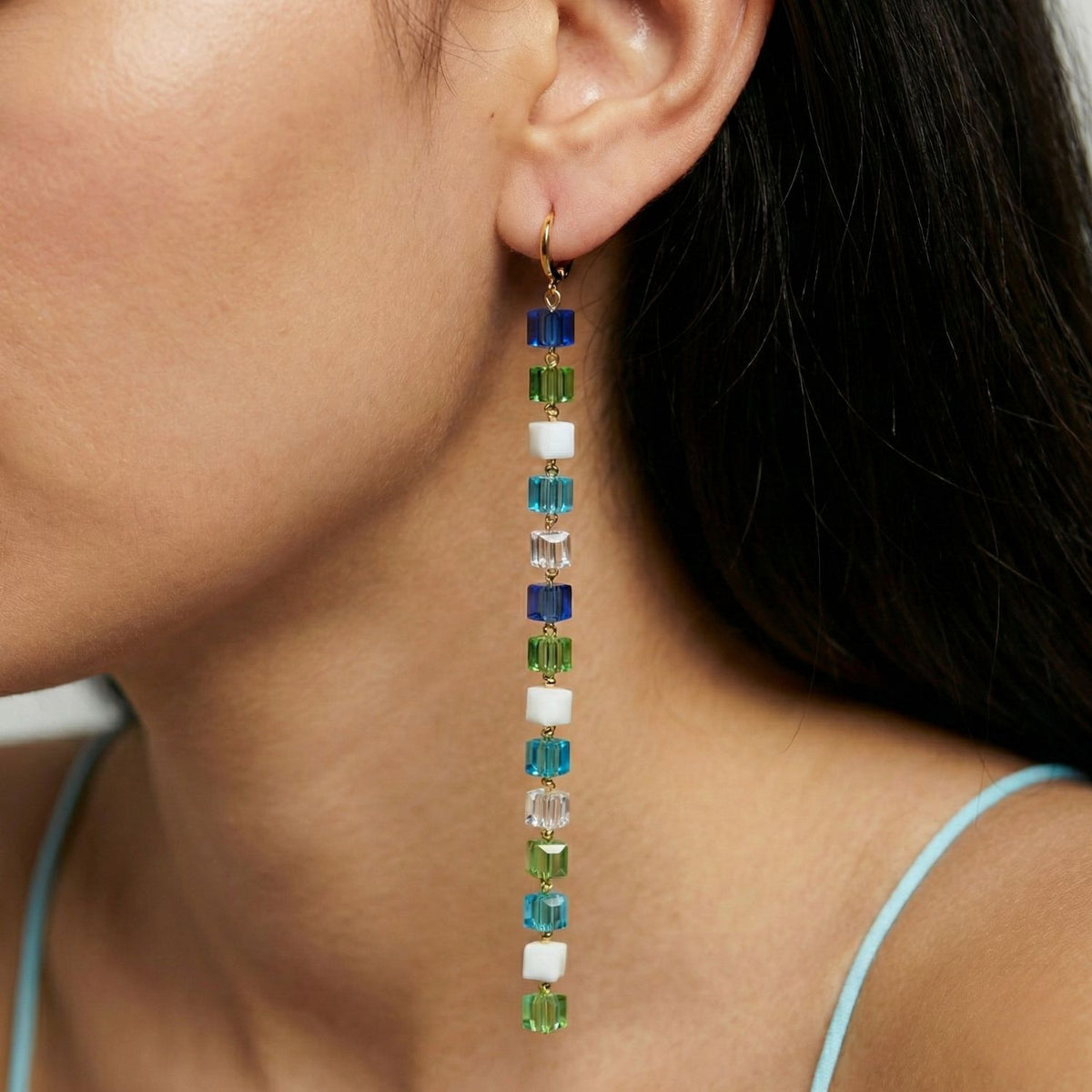 A woman with long dark hair wears JAREDJAMIN Jewelry Online's Blue Hue Building Block Drop Earrings, featuring stacked blue, green, and white glass beads on an 18K gold plated hook, paired with a light blue spaghetti strap top.