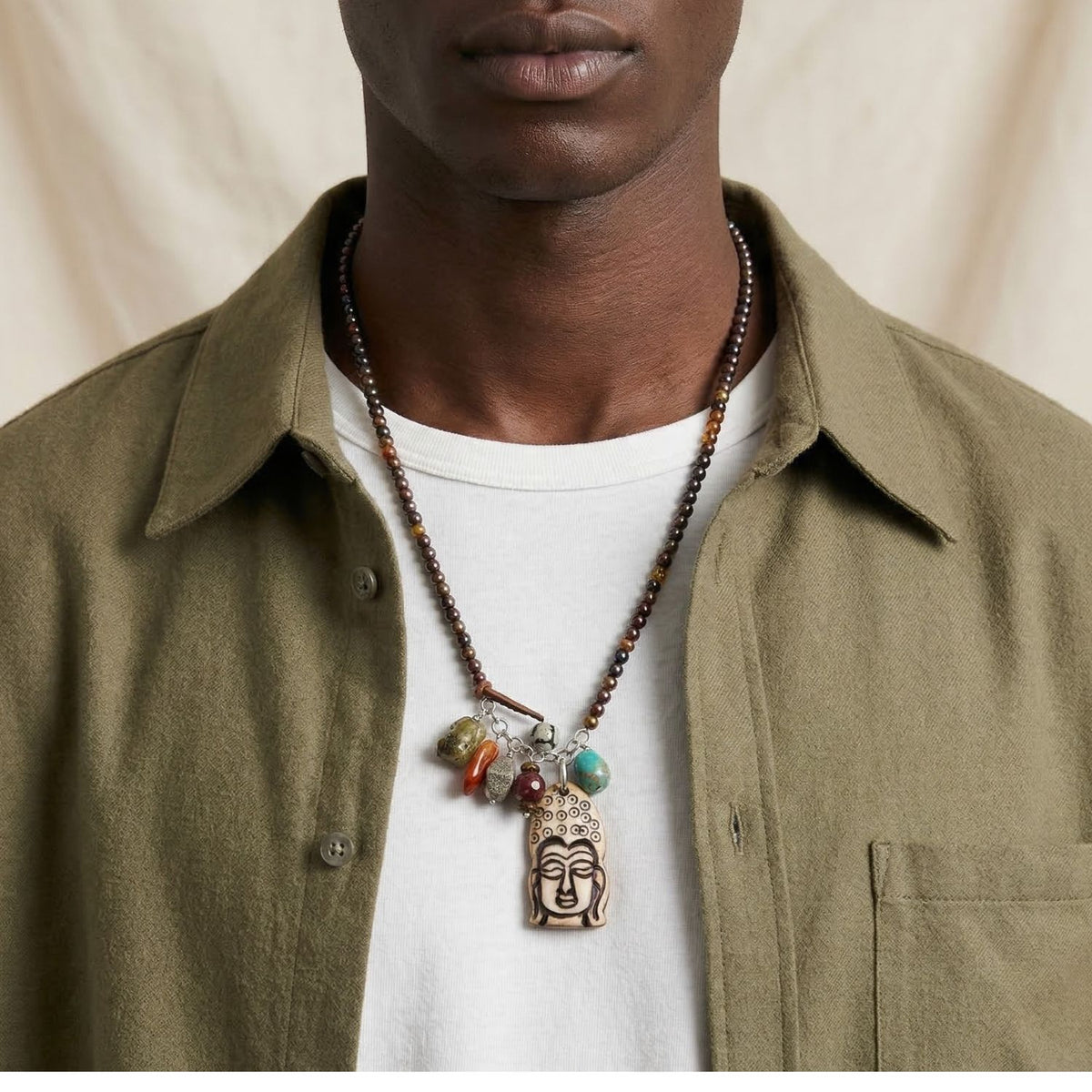 Los Buddha Meanderings Beaded collar de JAREDJAMIN Joyería Online features a large carved Buddha colgante y vibrant stone dijes, displayed against a softly lit neutral fabric background by a person in an olive overshirt y white tee.