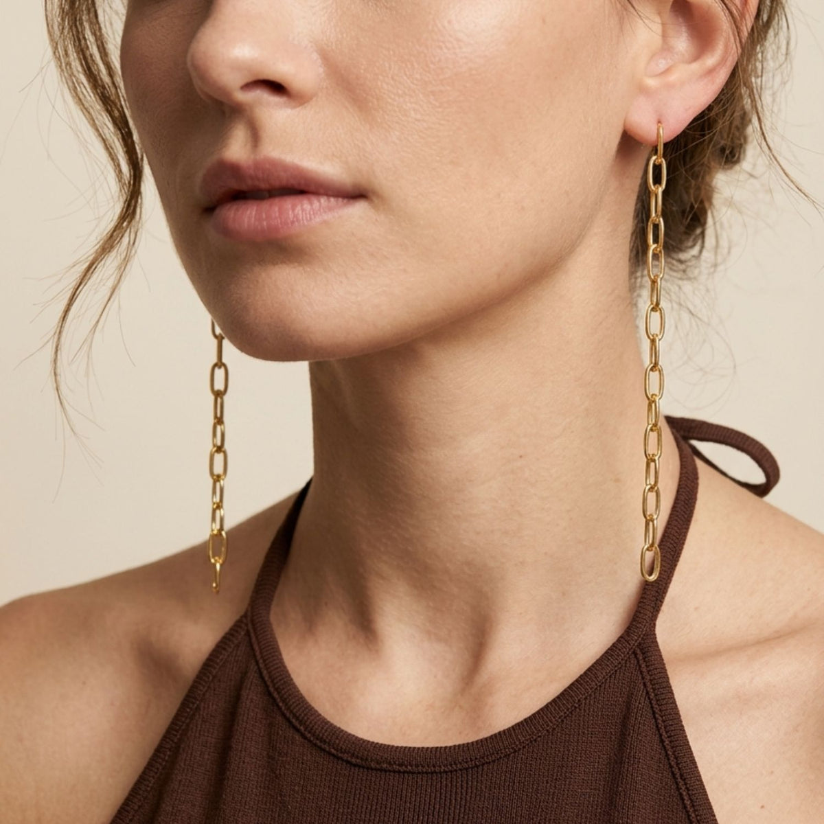 Side profile portrait view of a model wearing the 4.25-inch Chain Reaction Linear Link Earrings, demonstrating their fluid drape, architectural jawline-framing volume, and reflective gold finish styled with a brown halter top.