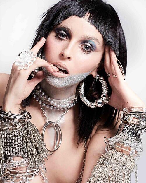 A person with short black hair and bold makeup poses with their hands near their face. They wear heavy silver and crystal jewelry, including large hoop earrings, necklaces, and numerous bracelets. A silver glitter pattern is painted across their chin and mouth.