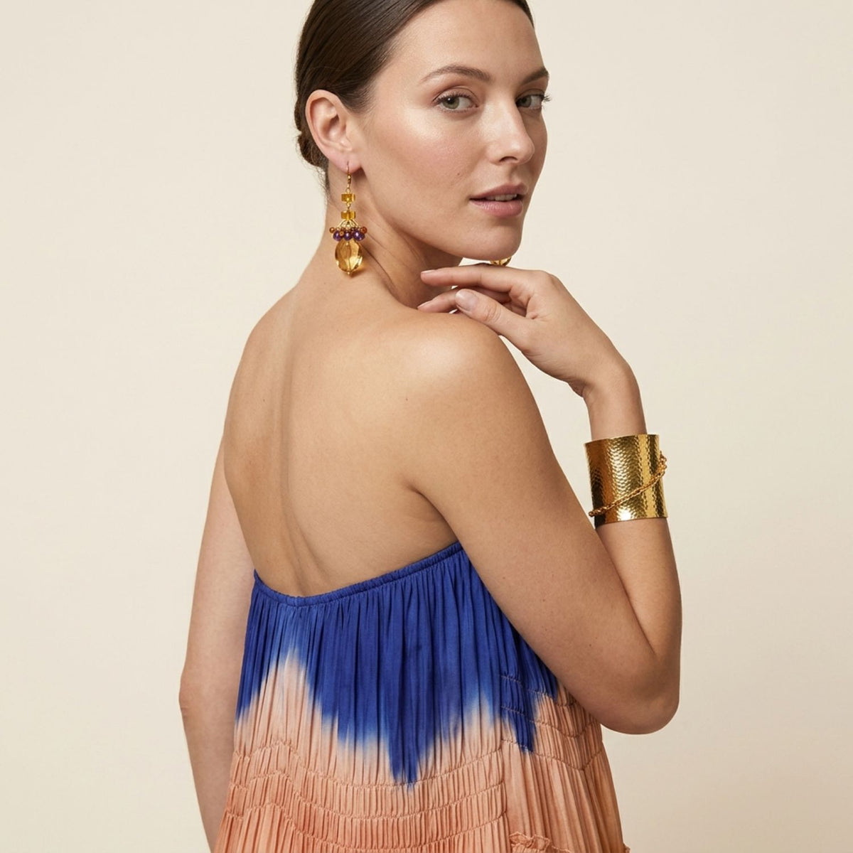 Over-the-shoulder view of a model wearing the Copacabana Amber Crystal and Amethyst earrings, styled elegantly with a blue and peach strapless tie-dye dress and a bold gold wrist cuff.