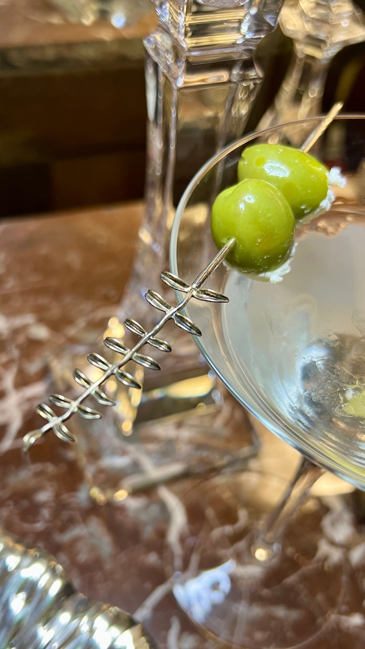 A martini glass with a clear cocktail and two green olives on a Silver Thyme skewer by JAREDJAMIN Barware sits elegantly against crystal objects and a brown marble pattern, creating a sophisticated setup that enhances any charcuterie board or party supplies collection.