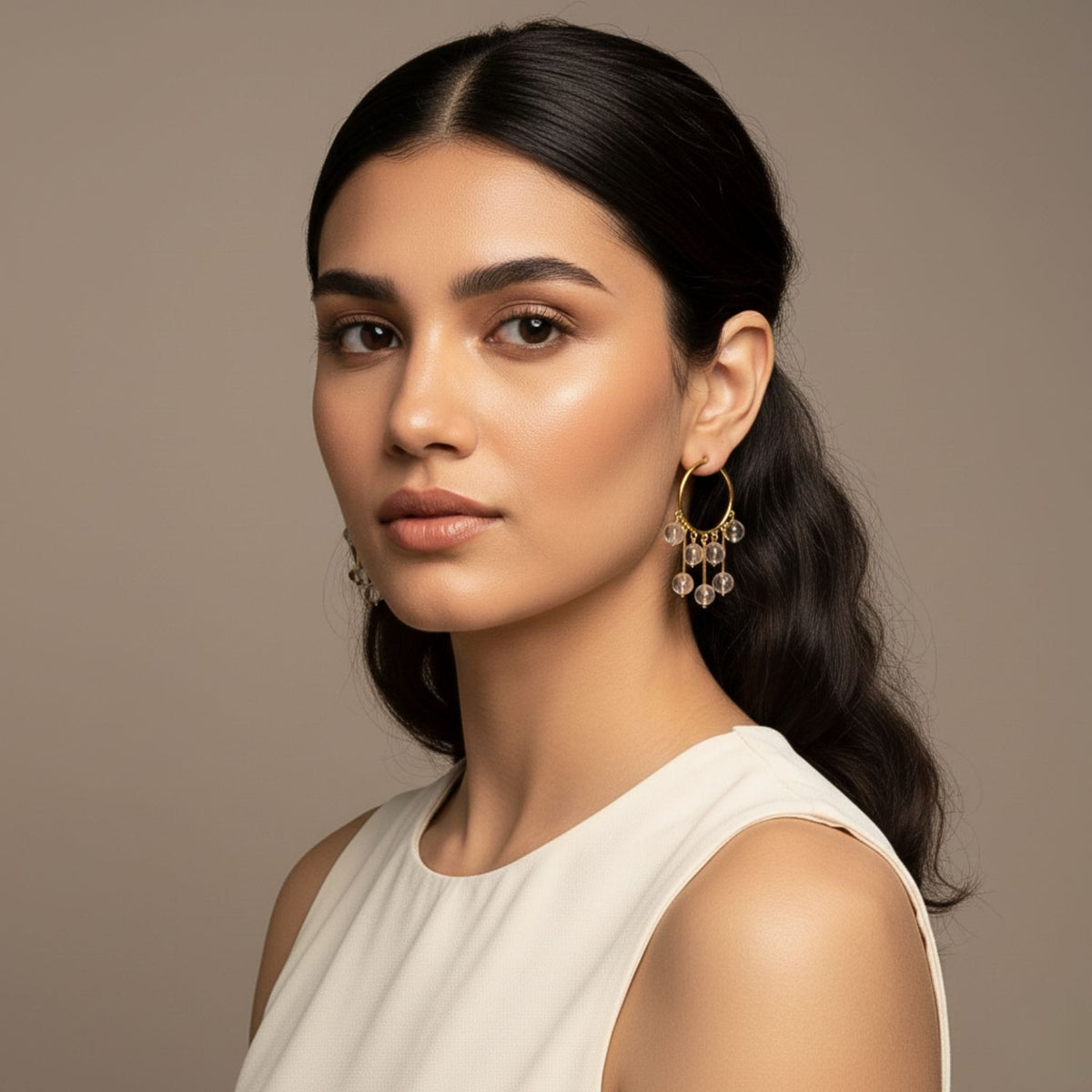 Un woman avec long dark hair wears a sleeveless cream top et Daniela Dangle créole Quartz boucles d'oreilles par JAREDJAMIN Bijoux en Ligne. She has glowing skin, subtle makeup, et looks confidently at the camera against a neutral background.