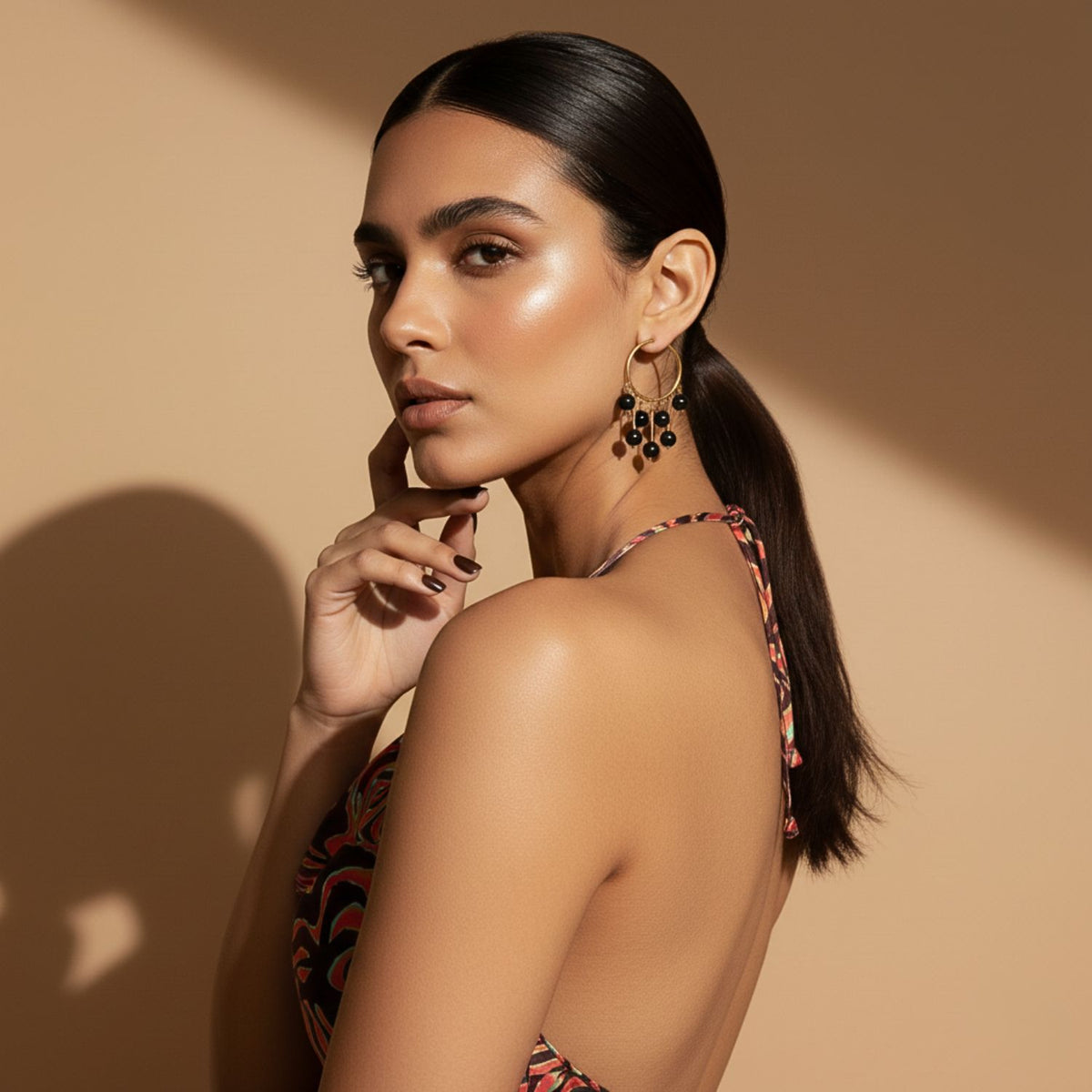 A woman with radiant skin and sleek dark hair in a low ponytail poses against a beige background, wearing Daniela Dangle Onyx Earrings from JAREDJAMIN Jewelry Online, a patterned halter top, and gazing confidently at the camera.