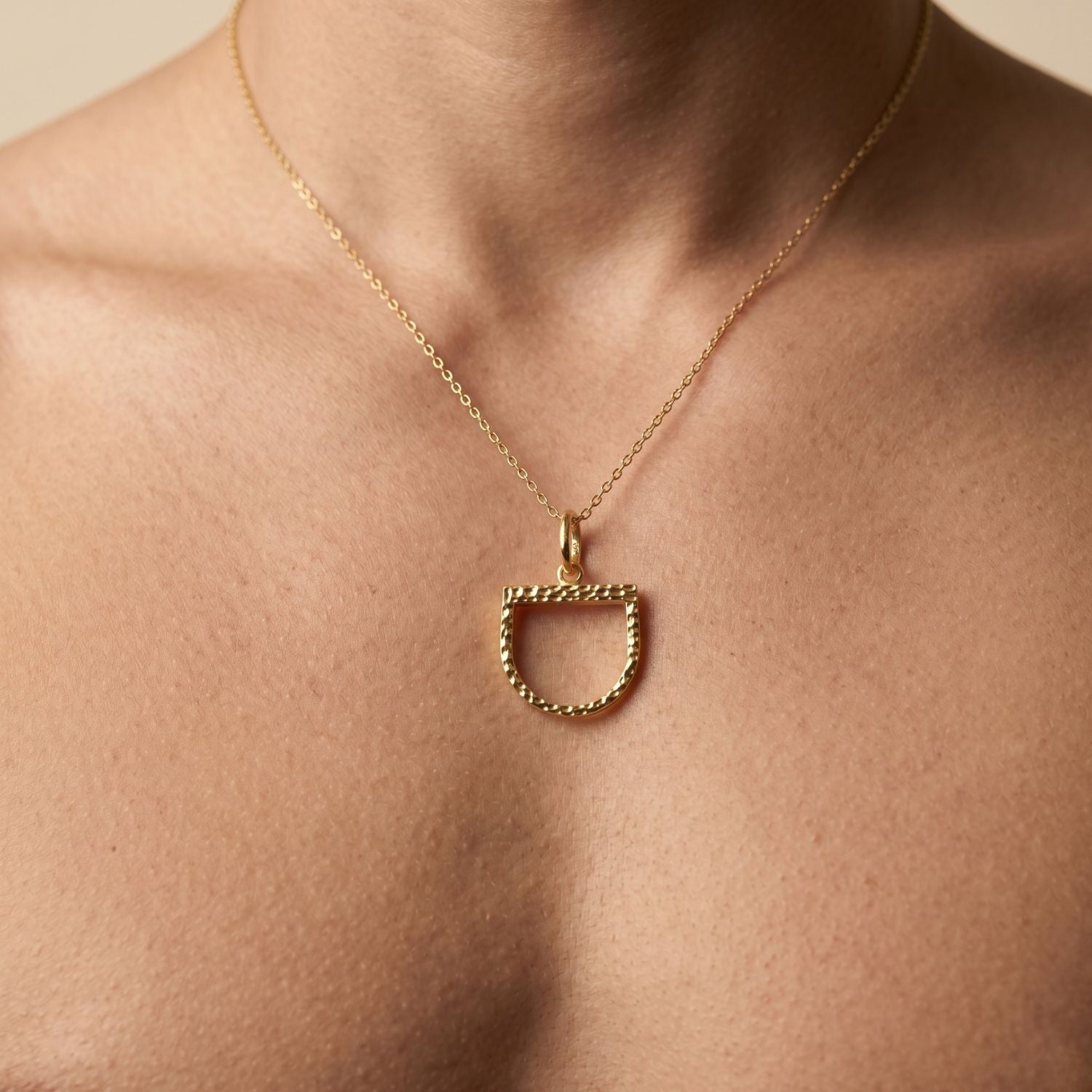 Minimalist studio shot of the JAREDJAMIN Darcy Pendant Charm against a solid background, showcasing the open-frame, stirrup-shaped "D" silhouette and its signature 18k gold-plated hammered texture suspended from a polished top jump ring.