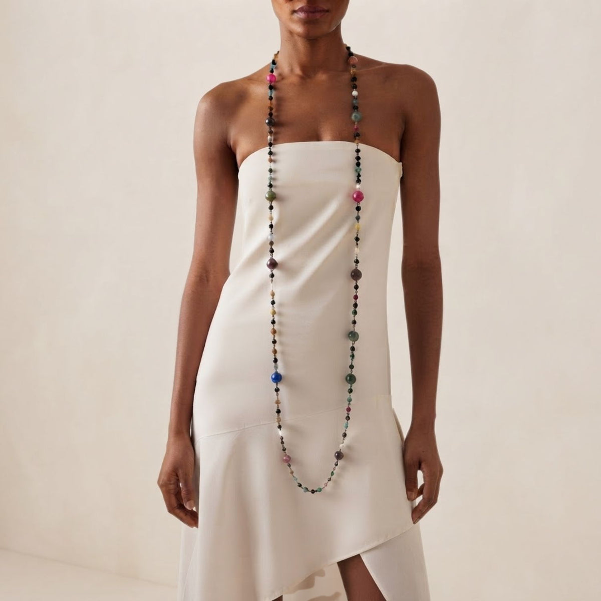 Front view of a model wearing the 70-inch gunmetal JAREDJAMIN Caelum Cosmic Convertible Strand long, showing its dramatic vertical line and colorful agate, onyx, and jasper planet beads against a strapless cream dress.
