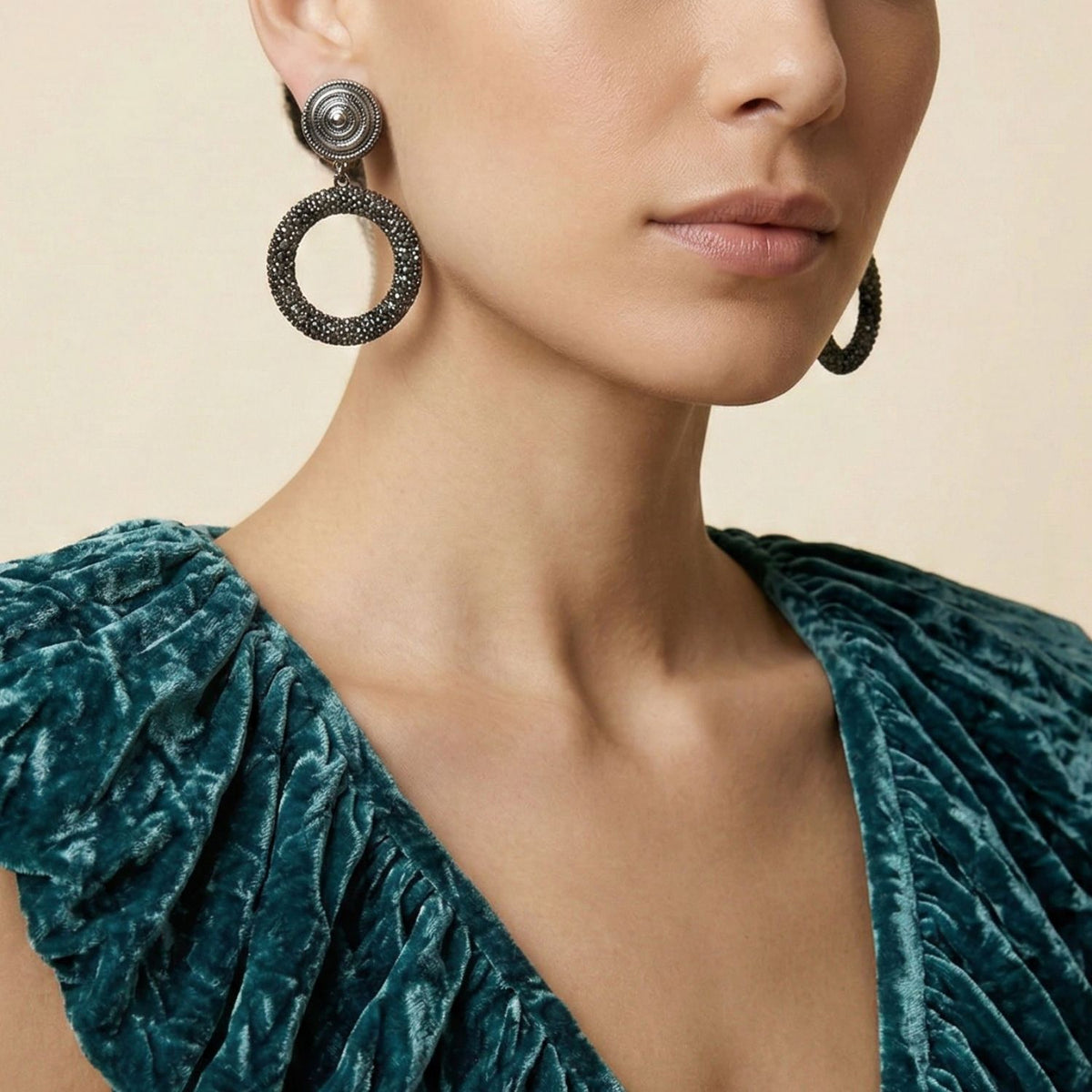 A woman in a teal velvet top with ruched sleeves is shown from the shoulders up, wearing JAREDJAMIN Jewelry Online's Dolly Pavé Clip On Earrings—statement earrings with textured studs and dark open hoops. Her expression is neutral.