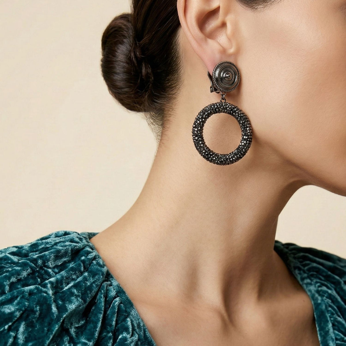 A woman with smooth, tan skin wears a textured teal velvet top and JAREDJAMIN Jewelry Online's Dolly Pavé Clip On Earrings. Her dark hair is styled in a low bun. The image shows her jaw to upper chest, facing right.