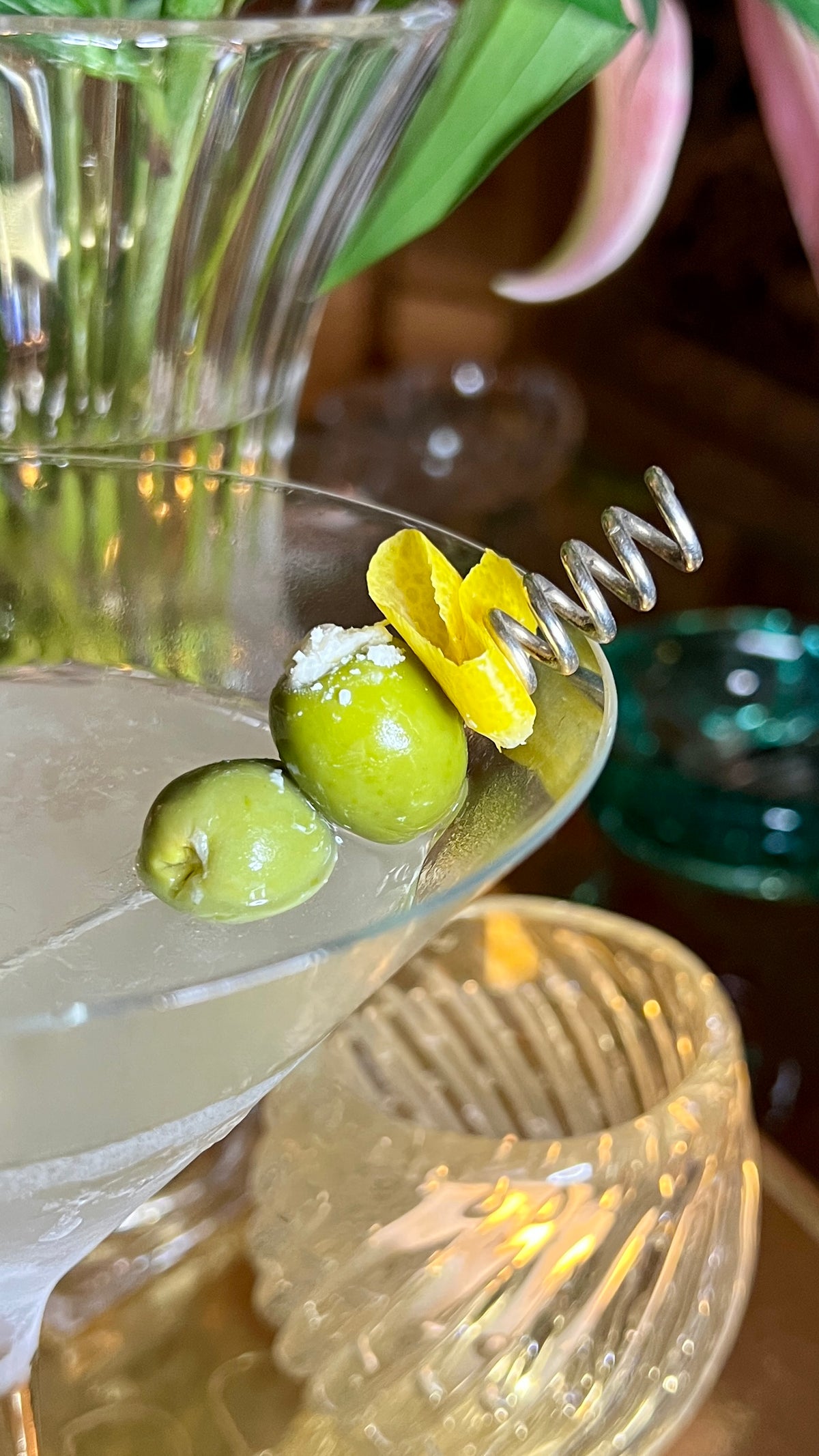 A close-up of a cocktail in a martini glass, garnished with two green olives on JAREDJAMIN Barware and More&#39;s Silver Corkscrew Cocktail Skewers, enhanced by elegant glassware, soft lighting, and sophisticated silver-plated party supplies.