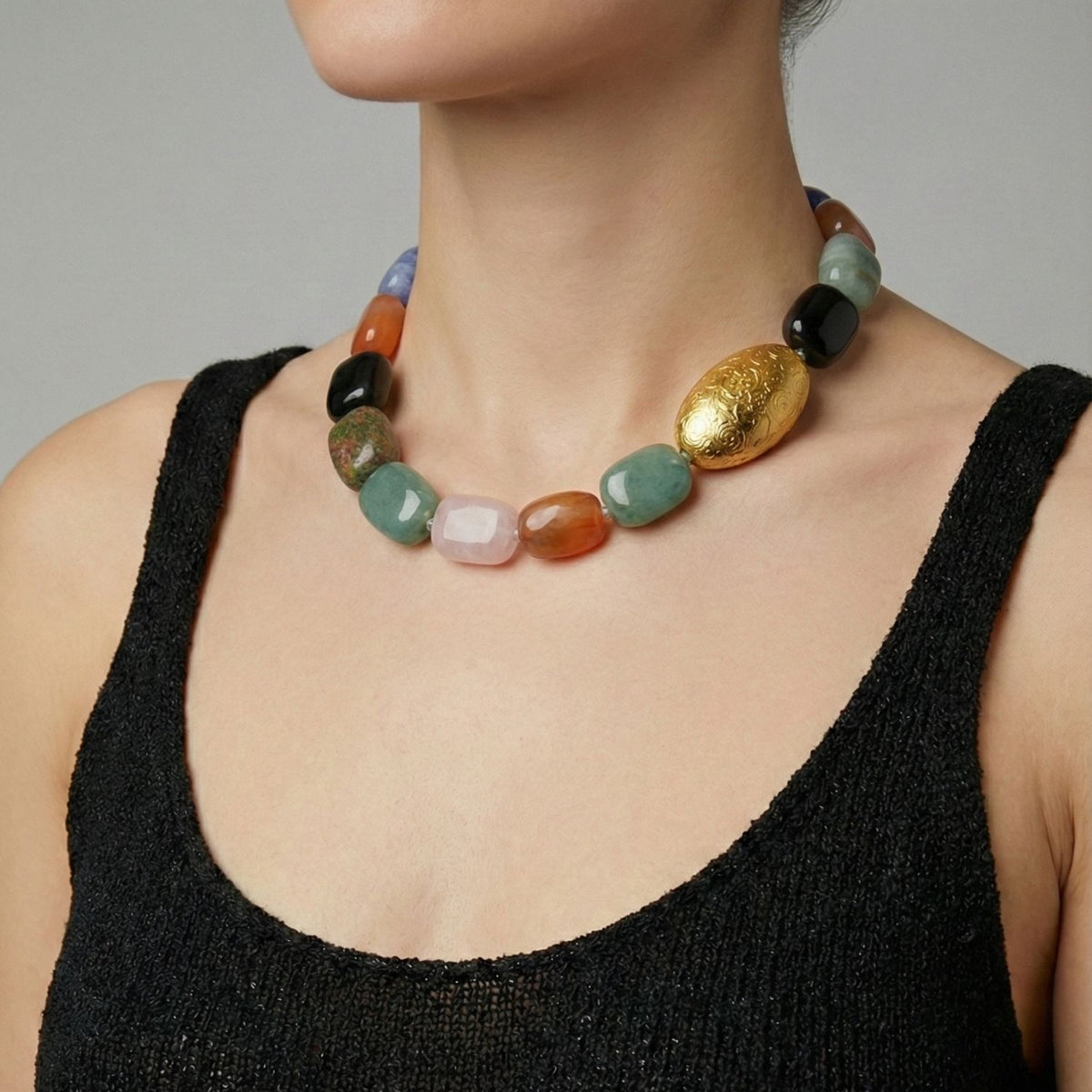 A woman in a sleeveless black textured top models the Earthen Elegance Gemstone Necklace by JAREDJAMIN Jewelry Online, featuring large multicolored gemstone beads and a striking oval engraved gold bead. Image is cropped below her nose.