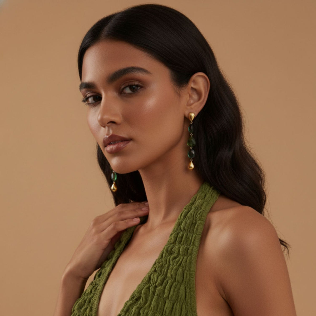 A womanとlong, wavy dark hair poses against a beige background. She wears a テクスチャード グリーン halter dressとJAREDJAMIN ジュエリーオンライン’s Emerald City イヤリング—ゴールド plated ステートメント piecesとグリーン bead accents. Her expression is calm.
