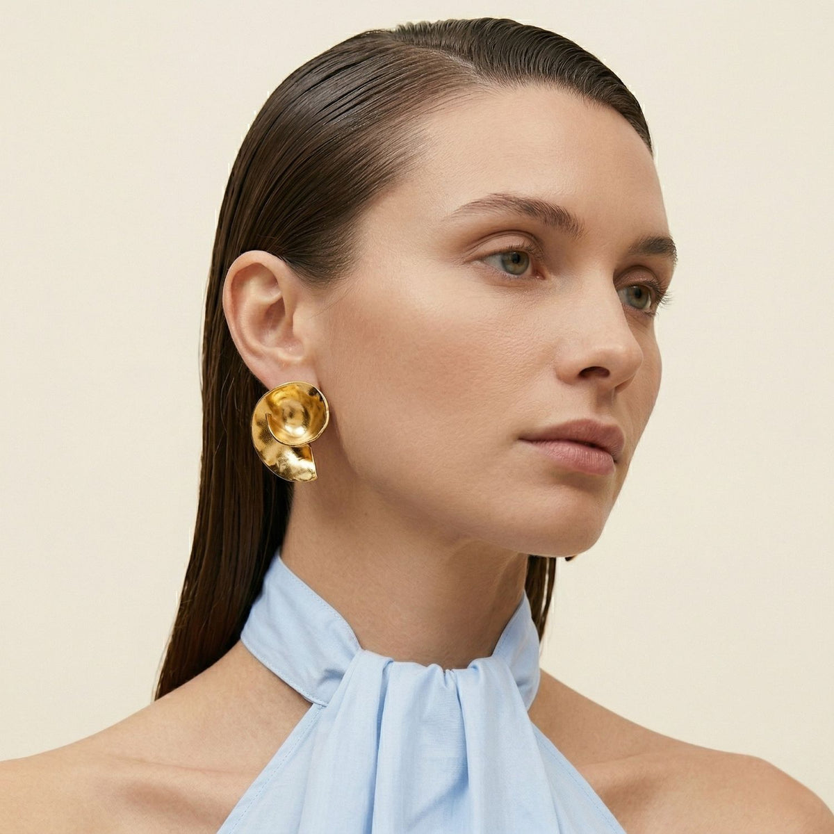 A woman with straight brown hair in a light blue halter top punctuates her look with hand-assembled Espiral Clip Earrings in 18k gold plate, crafted in our West Hollywood studio—small-batch, eco-conscious. Style Note: slips from workwear to evening edge.