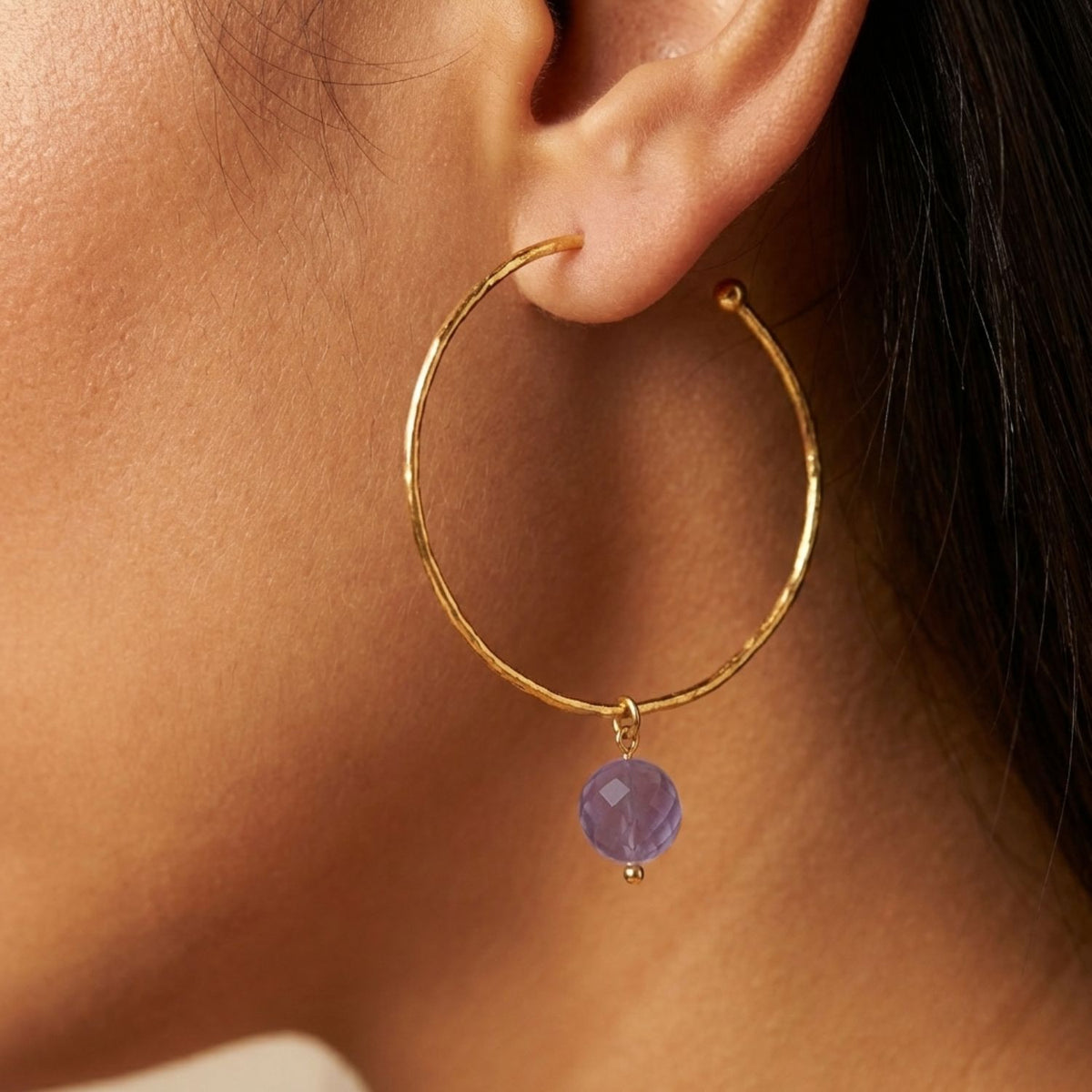 Un gros plan shows a femme portant a large, texturé 18K plaqué or créole de JAREDJAMIN Bijoux en Ligne, adorned avec a pendante lavender 16MM Faceted Ball boucle d'oreille breloque. Her dark hair is pulled back to highlight the élégant accessory.