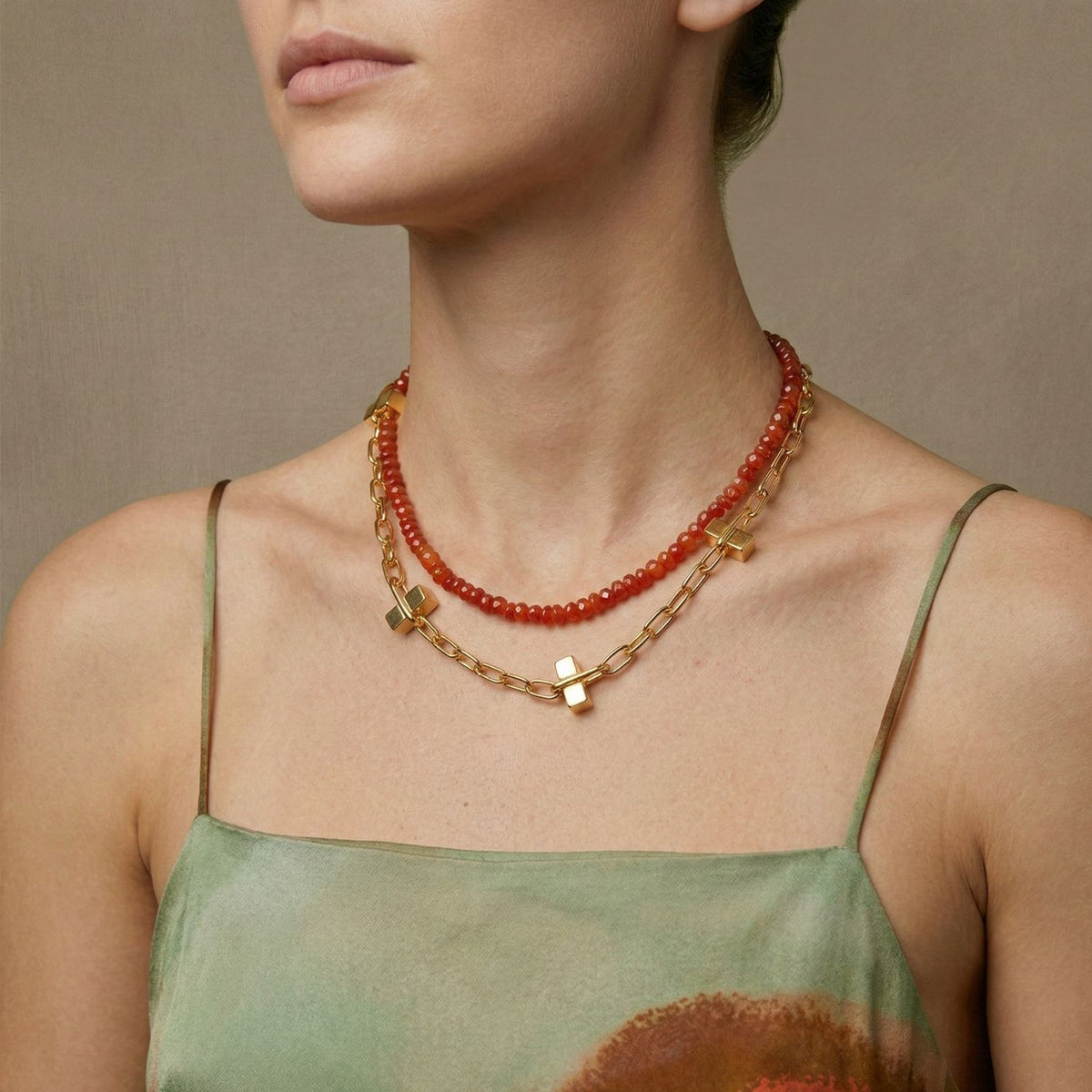 A woman in a green and brown dress wears two necklaces: the JAREDJAMIN Jewelry Online Gemstone Trail Carnelian Necklace, featuring vibrant carnelian roundelle beads, and a gold chain with square charms. A beige background highlights her elegance.