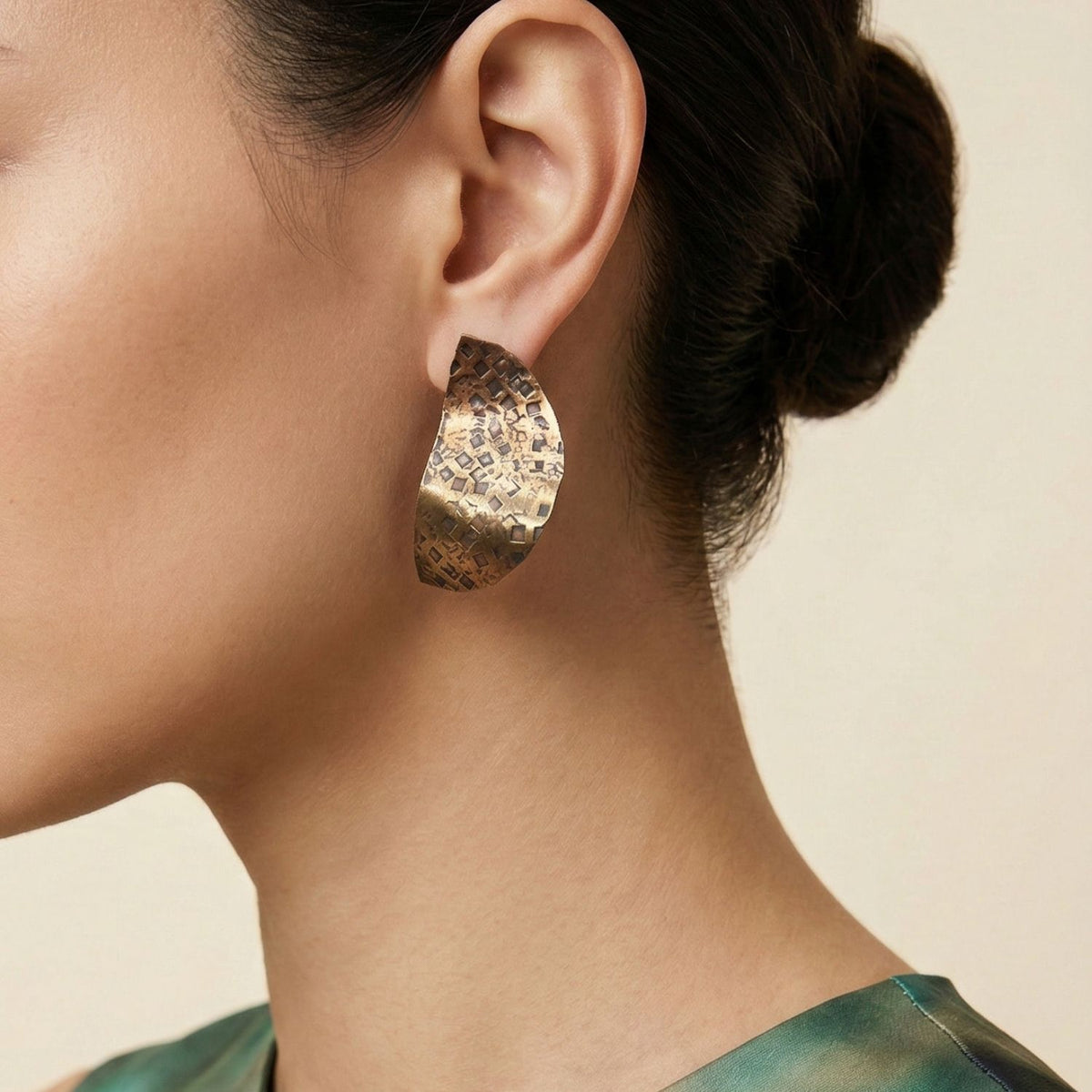 A close-up highlights a woman's head and neck as she wears JAREDJAMIN Jewelry Online’s Geo Twist Antique Tone Medium Earrings. Her dark hair is in a neat bun, and she pairs the earrings with a green satin-like top against a neutral background.