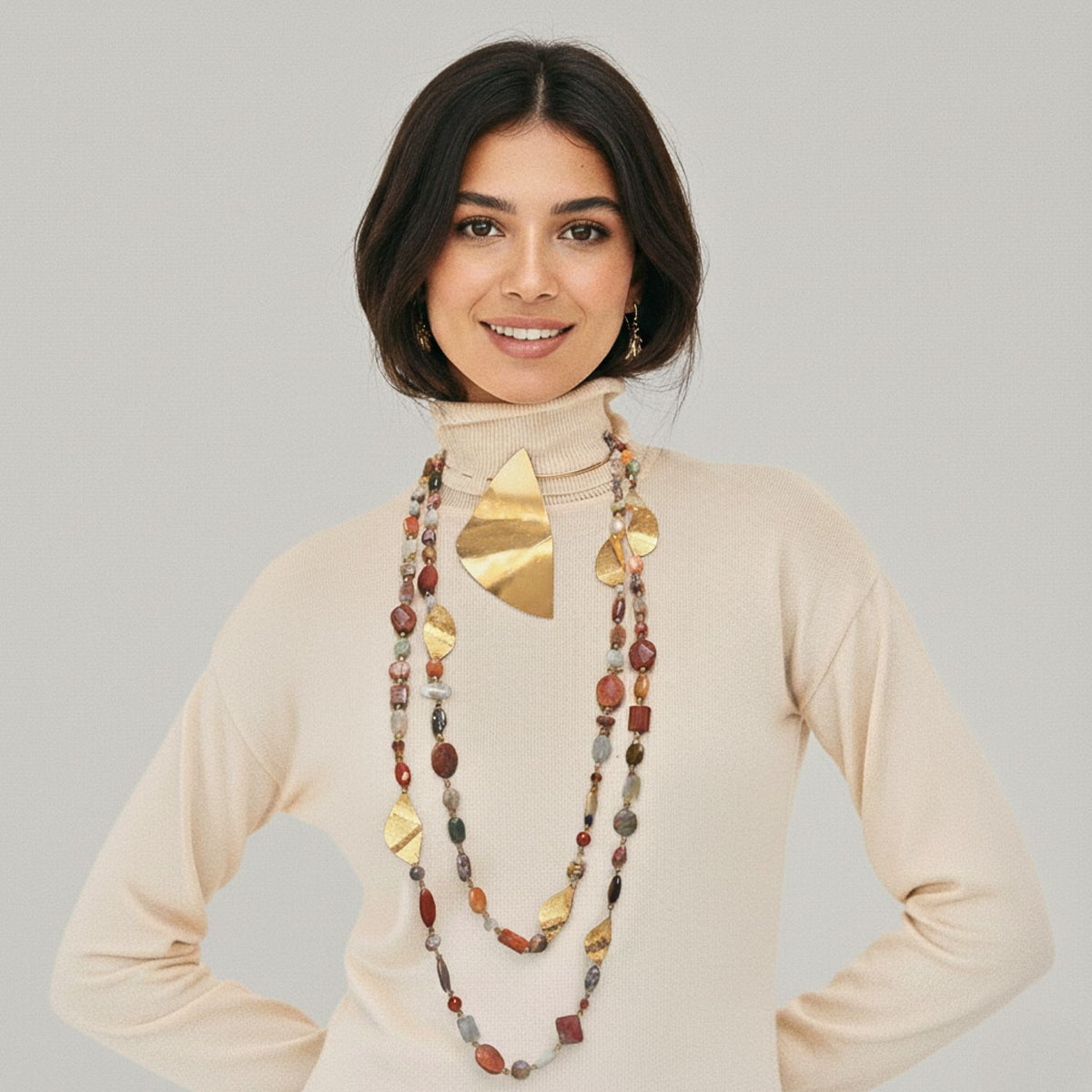 A woman with dark hair in a beige turtleneck sweater smiles, wearing the JAREDJAMIN Jewelry Online Geo Twist Gemstone Linked Necklace. She also wears a long hammered gold-plated charm necklace. The background is plain light gray.
