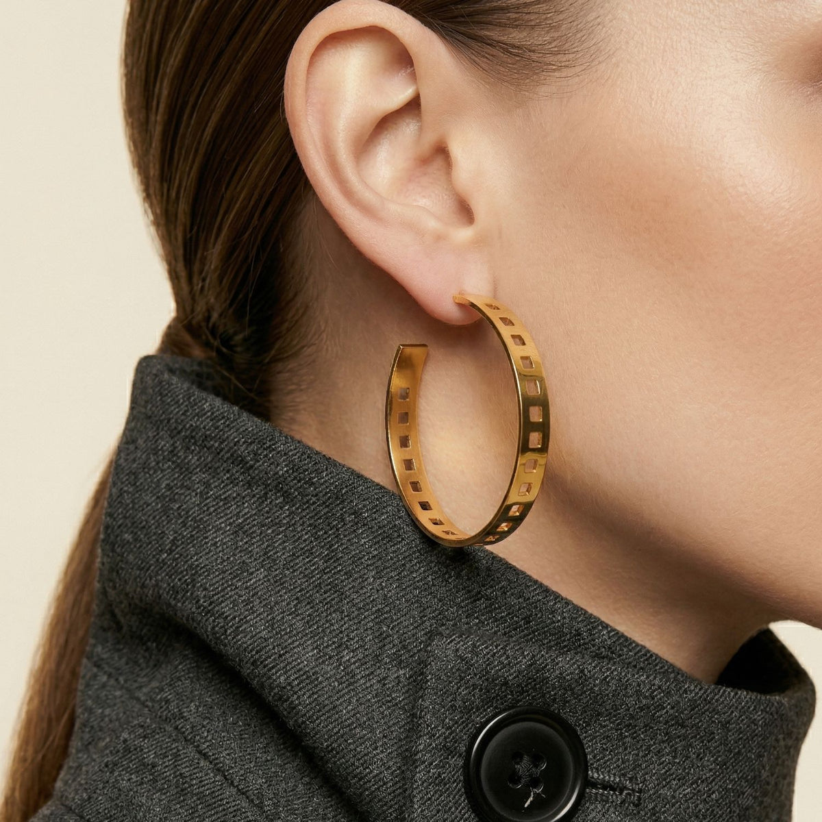 Close-up of a woman in a dark charcoal coat and black button, hair slicked back, punctuated by ‘Girls On Film’ Hoop Post Earrings—substantial gold arcs with geometric cut-outs, hand-assembled in West Hollywood using small-batch, eco-minded methods. Style note: shifts seamlessly from tailored suiting to after-dark silhouettes.