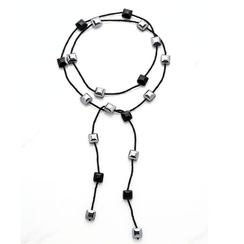 A black glass bead lariat necklace laid in a loose circle on a white background: black seed beads interspersed with faceted black and mirror-silver square crystals, with two long beaded tails.
