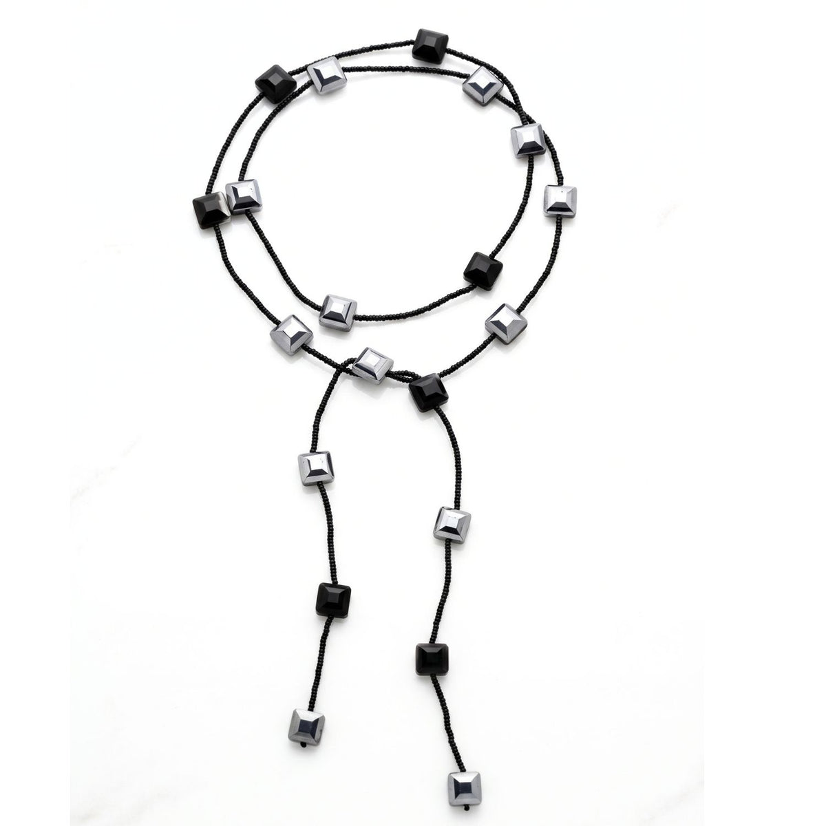 A black glass bead lariat necklace laid in a loose circle on a white background: black seed beads interspersed with faceted black and mirror-silver square crystals, with two long beaded tails.