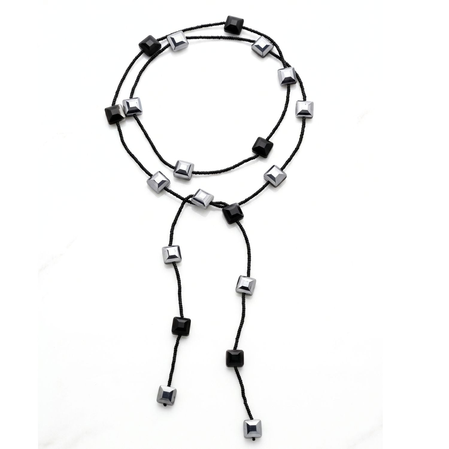 A black glass bead lariat necklace laid in a loose circle on a white background: black seed beads interspersed with faceted black and mirror-silver square crystals, with two long beaded tails.