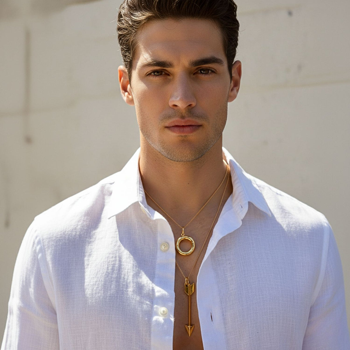 A young man with short dark hair wears a white, open button-down shirt and two layered chain necklaces, including the JAREDJAMIN Jewelry Online Gold Arrow Necklace Pendant Charm, against a sunlit backdrop.