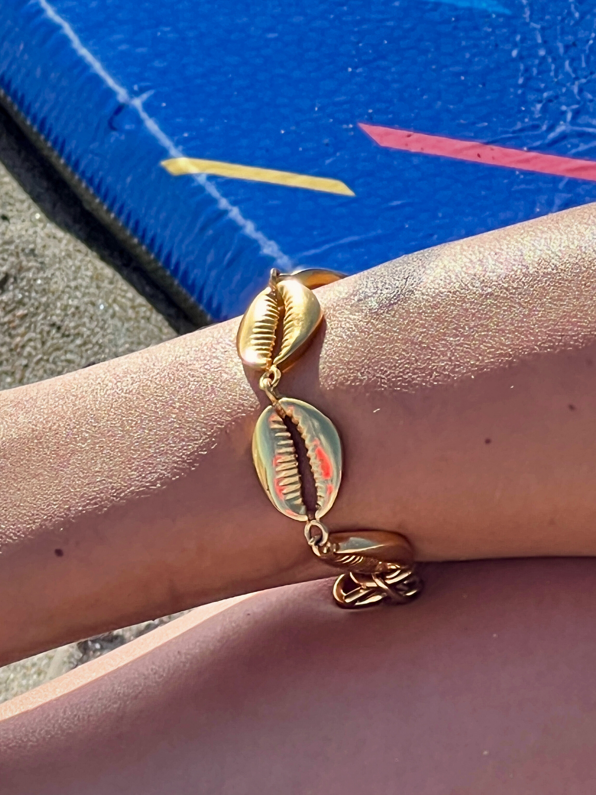 A close-up of an arm displays the Gold Bel Mare Cowrie Seashell Bracelet by JAREDJAMIN Jewelry Online. The skin glimmers in sunlight, accentuating the toggle clasp, as a colorful inflatable raft is blurred against a sandy backdrop.