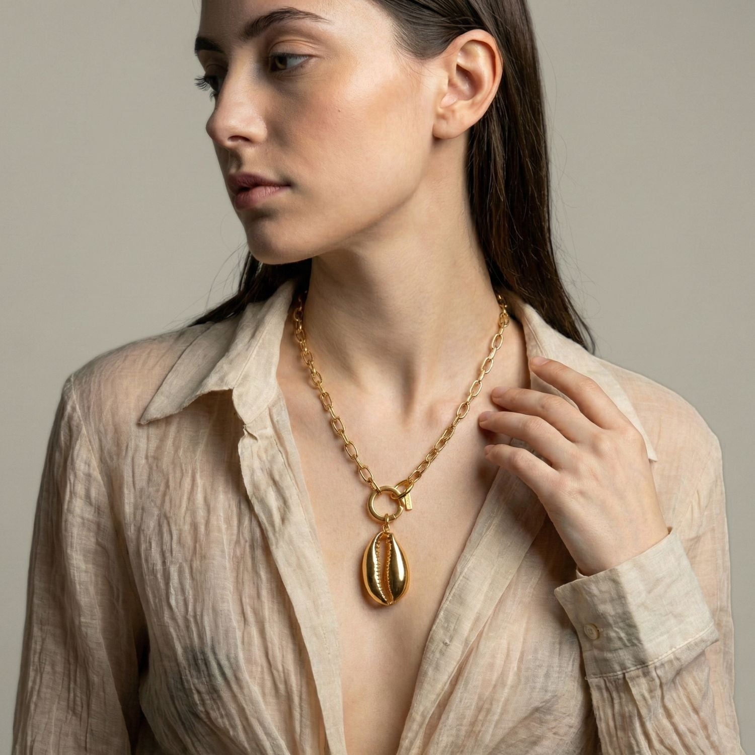 A Gold Bel Mare Cowrie Seashell Pendant Charm Chain Necklace by JAREDJAMIN Jewelry Online is showcased over a sheer beige blouse, with the model gazing left and her right hand gently touching the necklace against a neutral background.