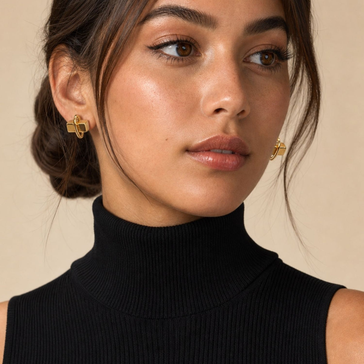 Close-up front portrait of a female model with dark hair wearing a black sleeveless top, styling the geometric Gold Blockbuster Studs to demonstrate how the 3D cube silhouette sits proudly against the earlobe.