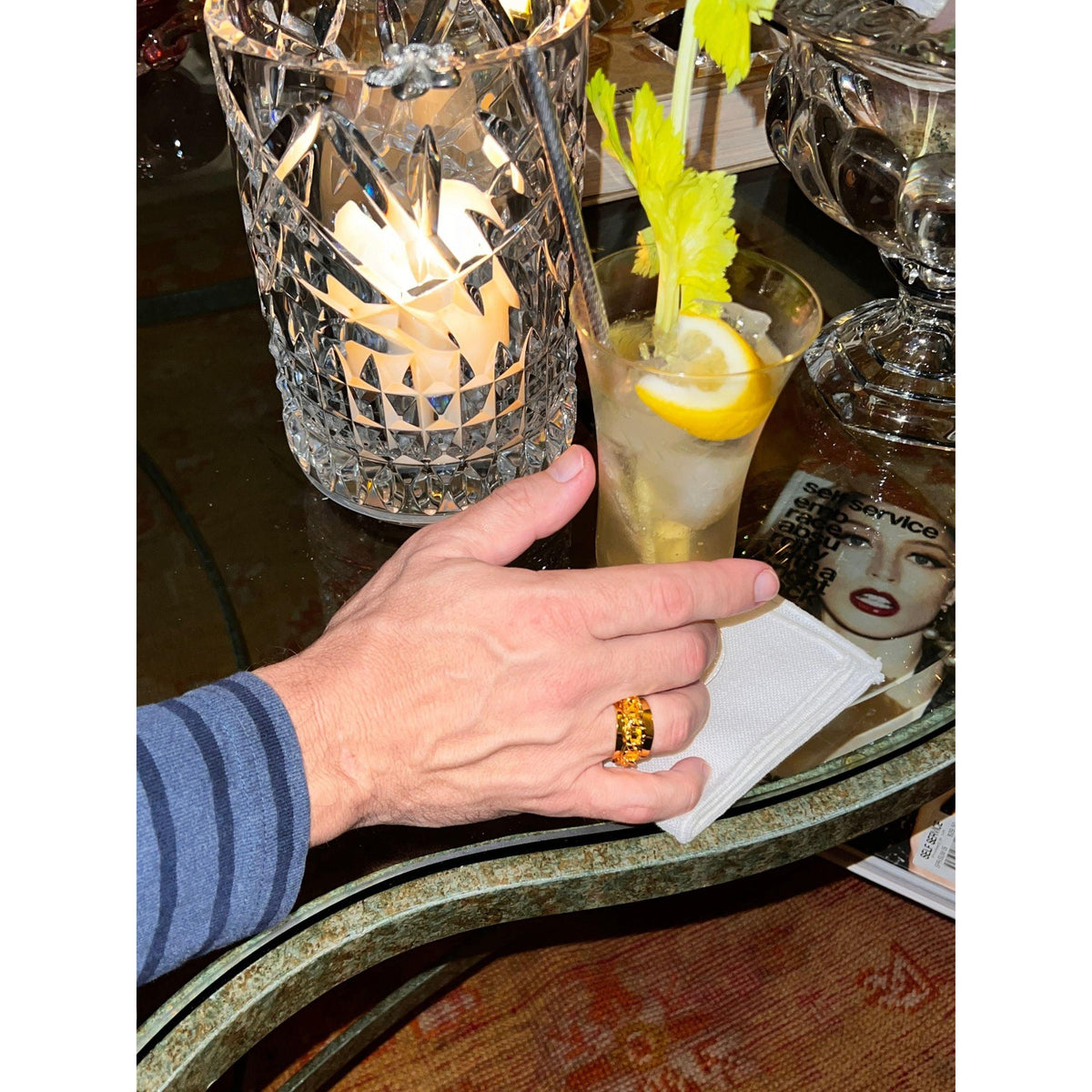 A hand wearing the JAREDJAMIN Jewelry Online Gold Charlie Chain Mens Ring reaches for a celery- and lemon-garnished drink in a tall glass on a napkin; nearby are a lit crystal candle holder, magazine, and glass bowl on the glass-top table.